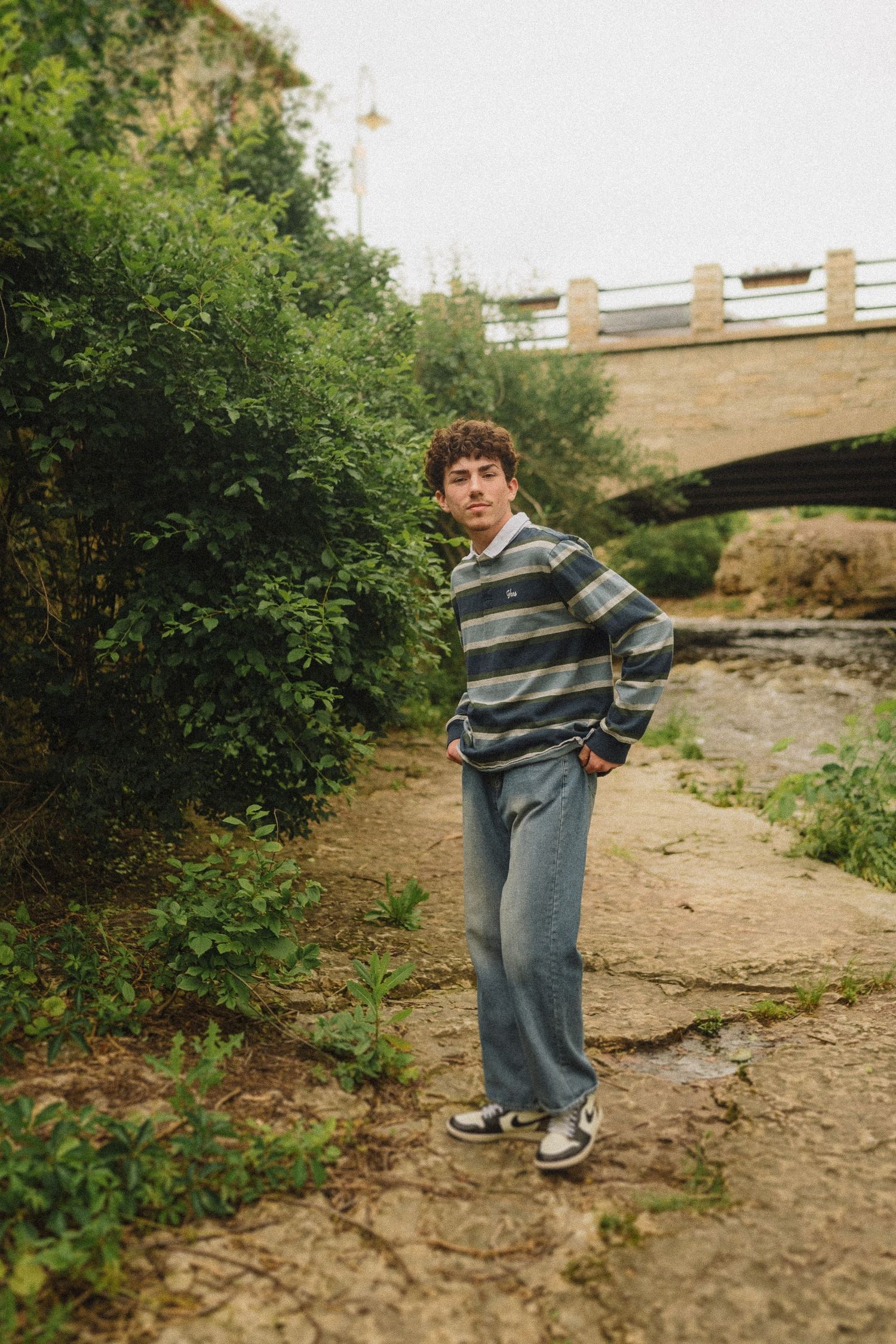 Someone wearing a striped sweater and jeans stands on a dirt path beneath a stone bridge in a wooded area.