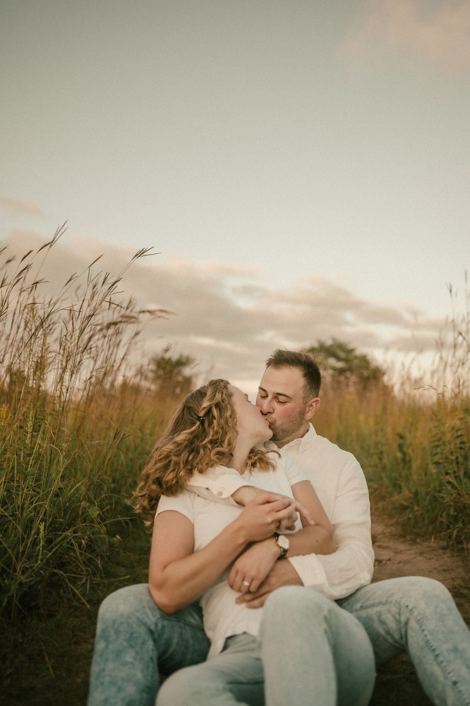 Romantic couple shares intimate moment while sitting in tall grass during golden hour.