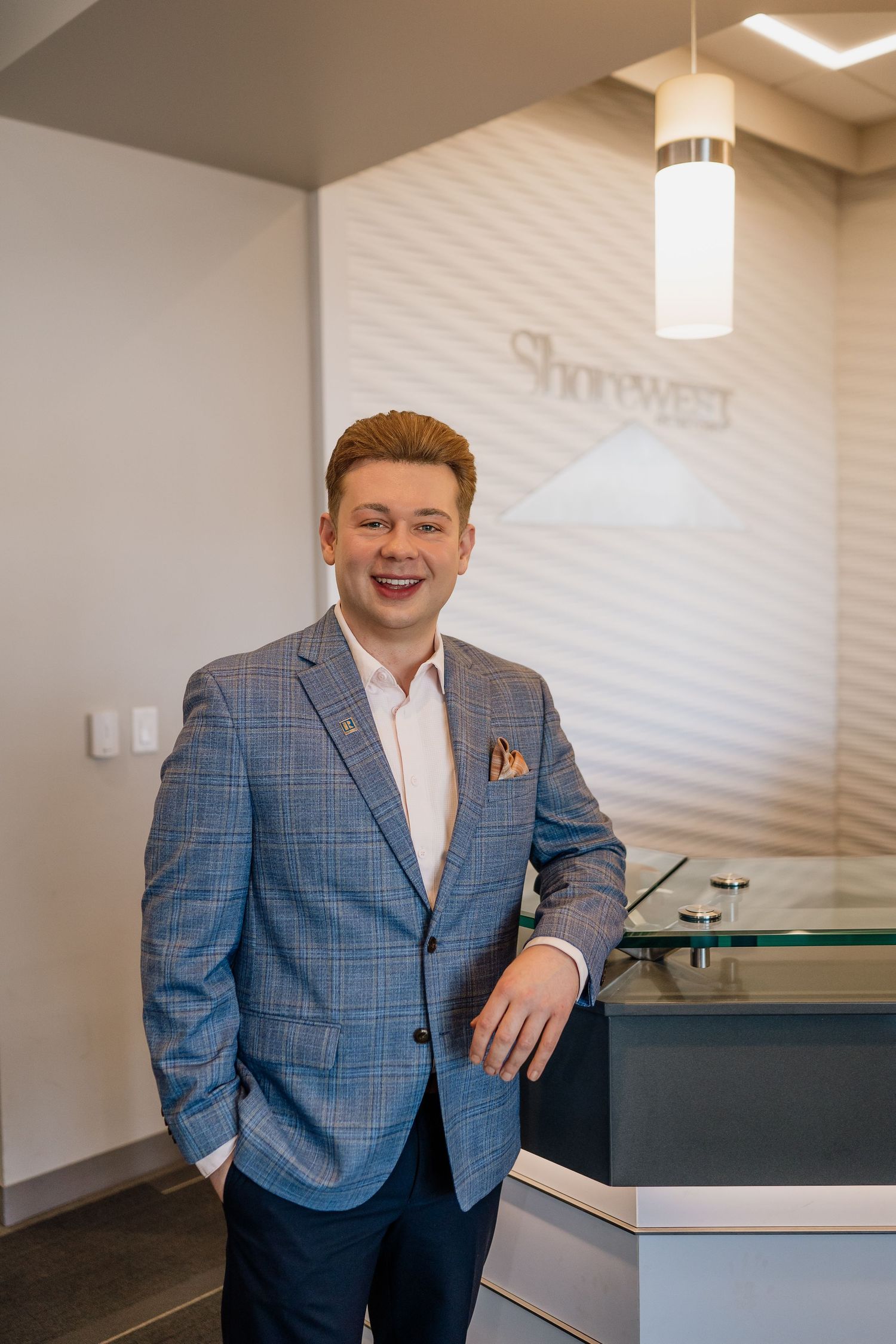 Business professional in blue checkered suit jacket poses in modern office space.