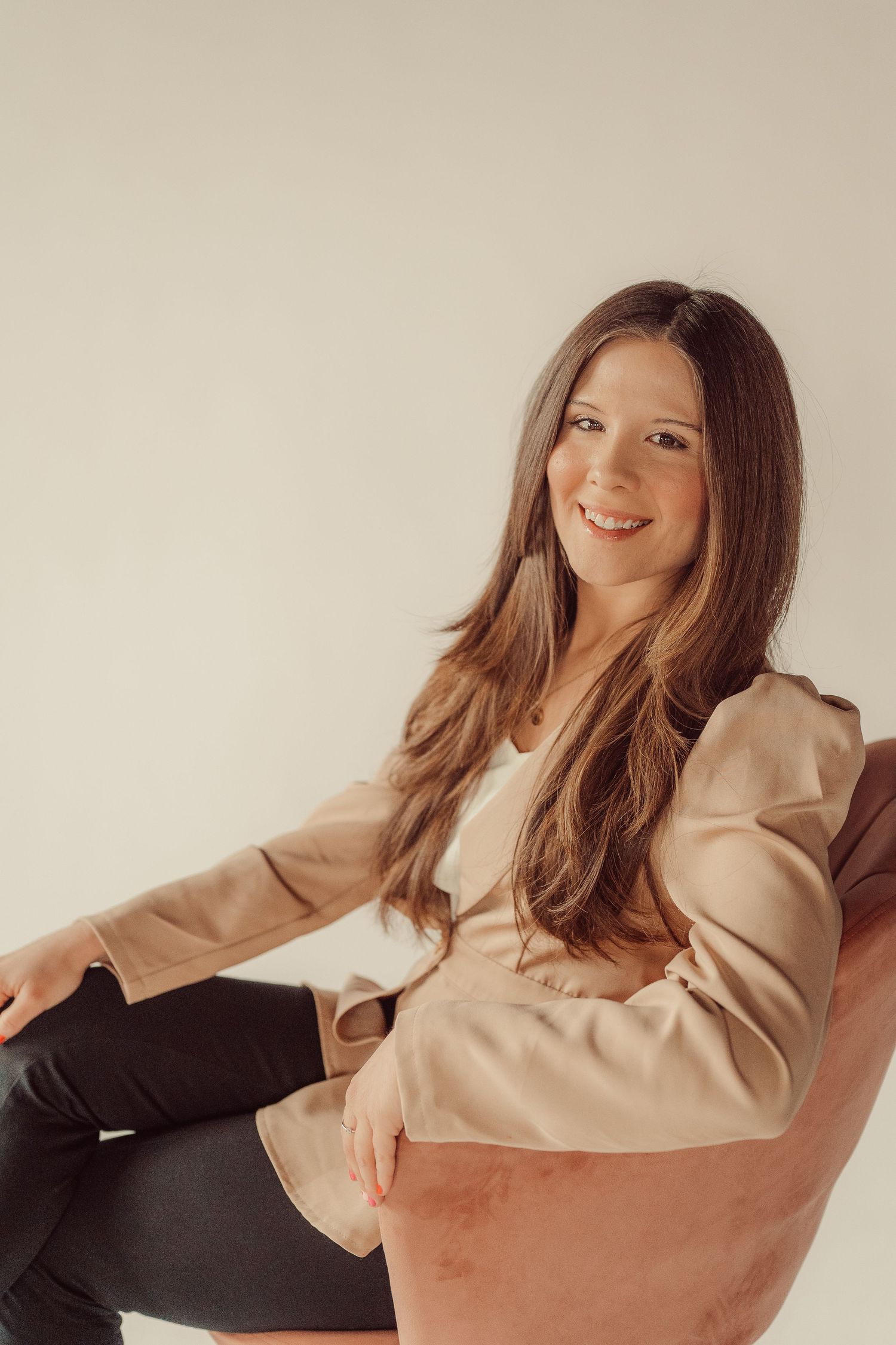 Casual portrait of woman seated in orange velvet chair wearing beige blazer and black pants.
