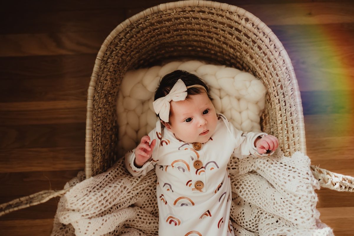 Baby with white bow rests in woven basket with knit blanket and rainbow light effect.