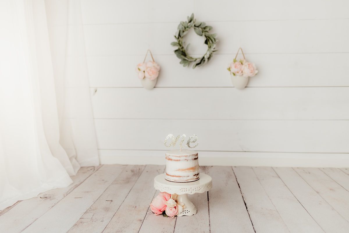 Minimalist cake smash setup with white cake and floral decorations against white wooden backdrop.