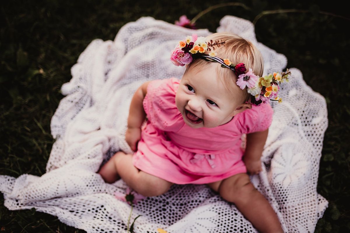 Baby in pink outfit and floral crown smiles while sitting on white lace blanket in outdoor setting.