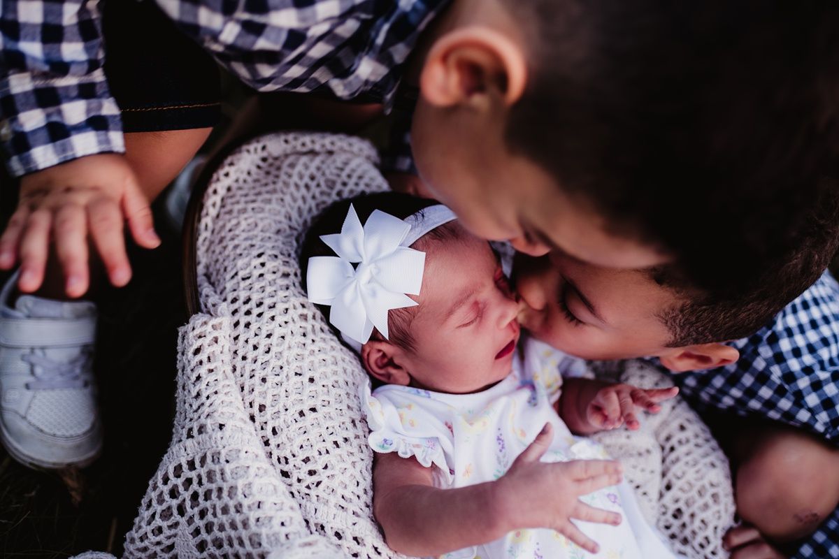 A newborn baby wearing a white bow lies wrapped in a crocheted blanket receiving a gentle kiss.