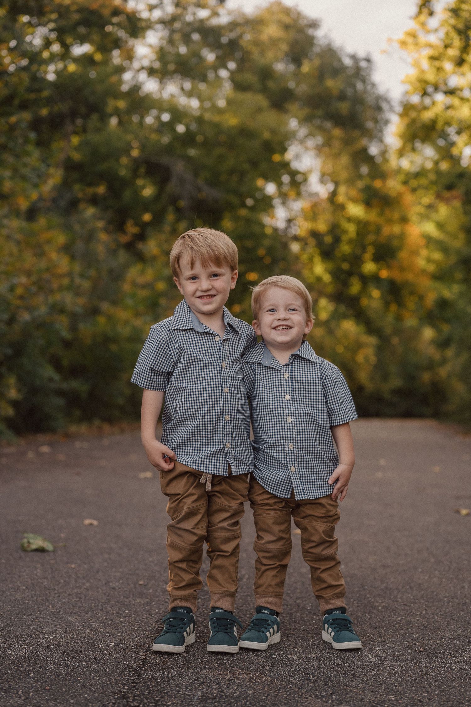 Two children in matching plaid shirts and brown pants stand together on a path with autumn trees in the background.