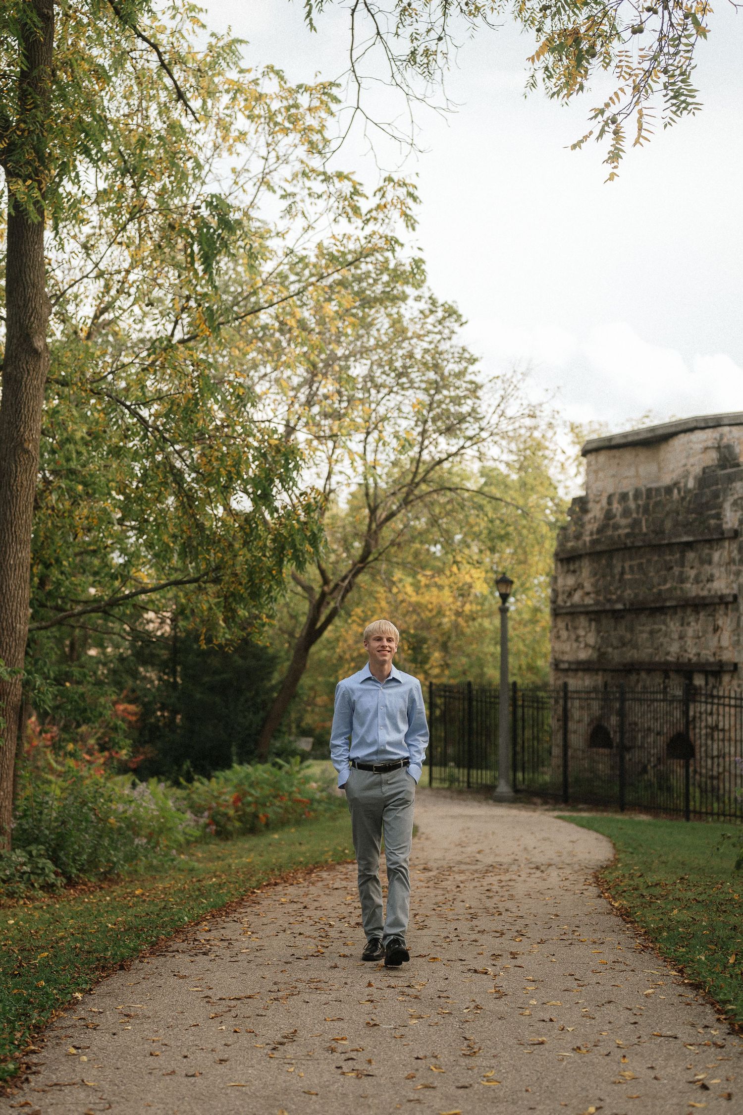 A person walks along an autumn path with trees and stone architecture in the background.