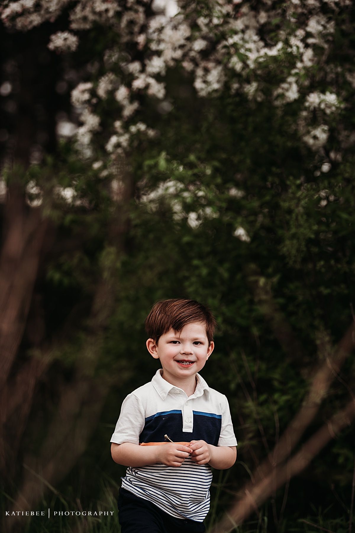 A young child in a striped shirt smiles brightly while standing under flowering trees.