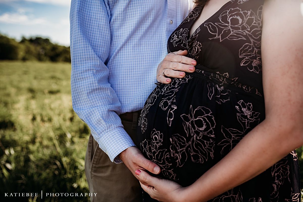 A couple embraces in a sunlit field as they capture a maternity moment in a floral dress and blue checkered shirt.