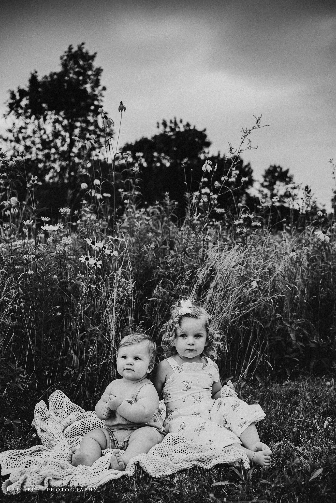 Black and white image of two small children sitting on a blanket in a wildflower meadow.