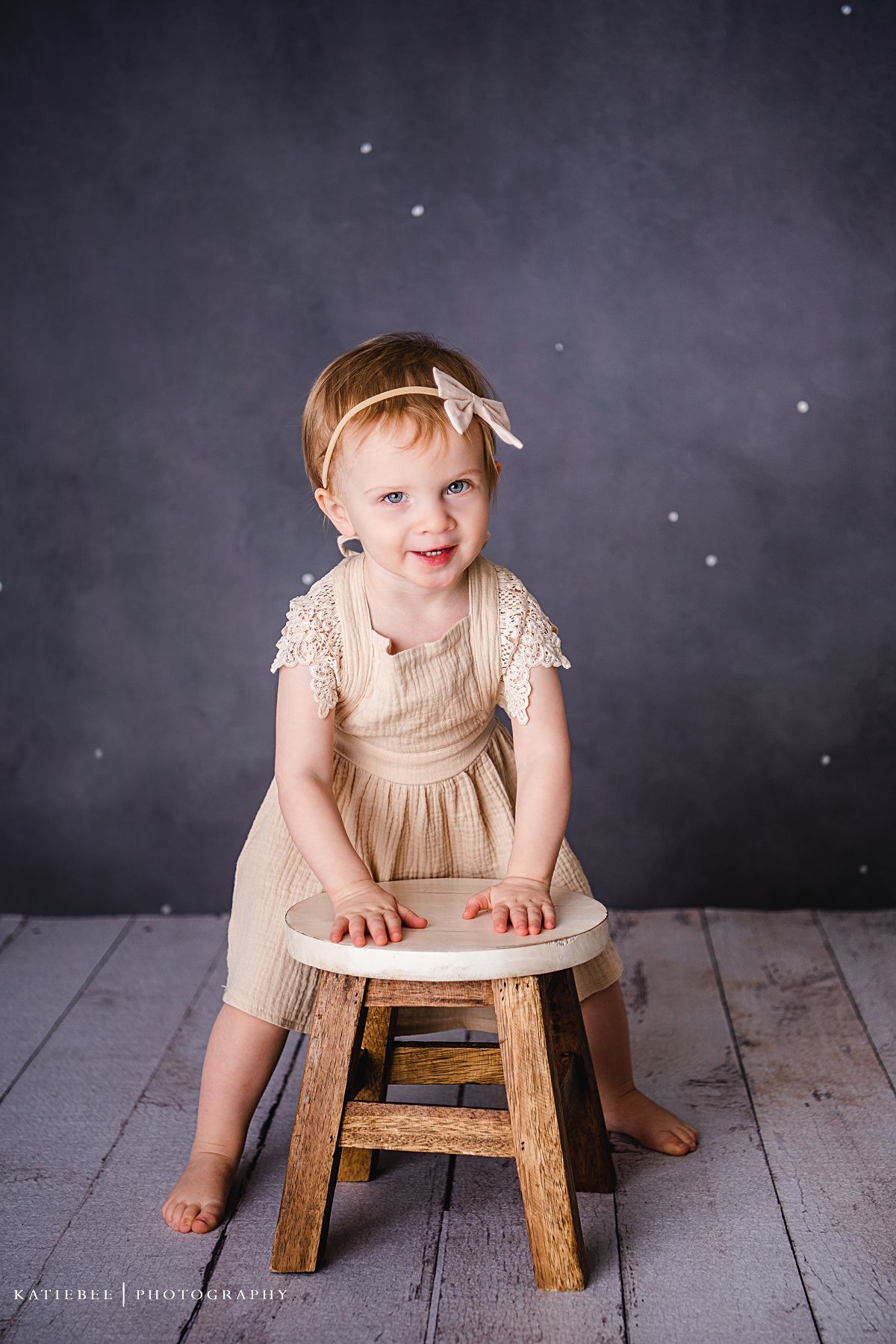 Child in lace dress sitting on wooden stool against dark sparkly background.