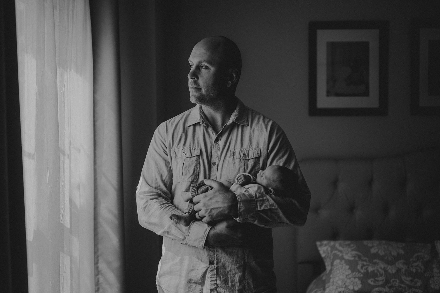 Parent in button-down shirt cradles sleeping infant near sunlit window.