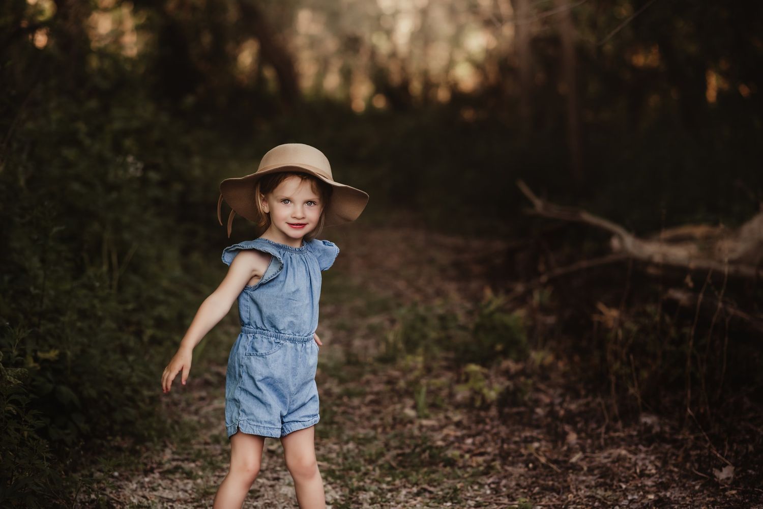 A denim romper and wide-brimmed hat outfit worn in a woodsy setting at sunset.