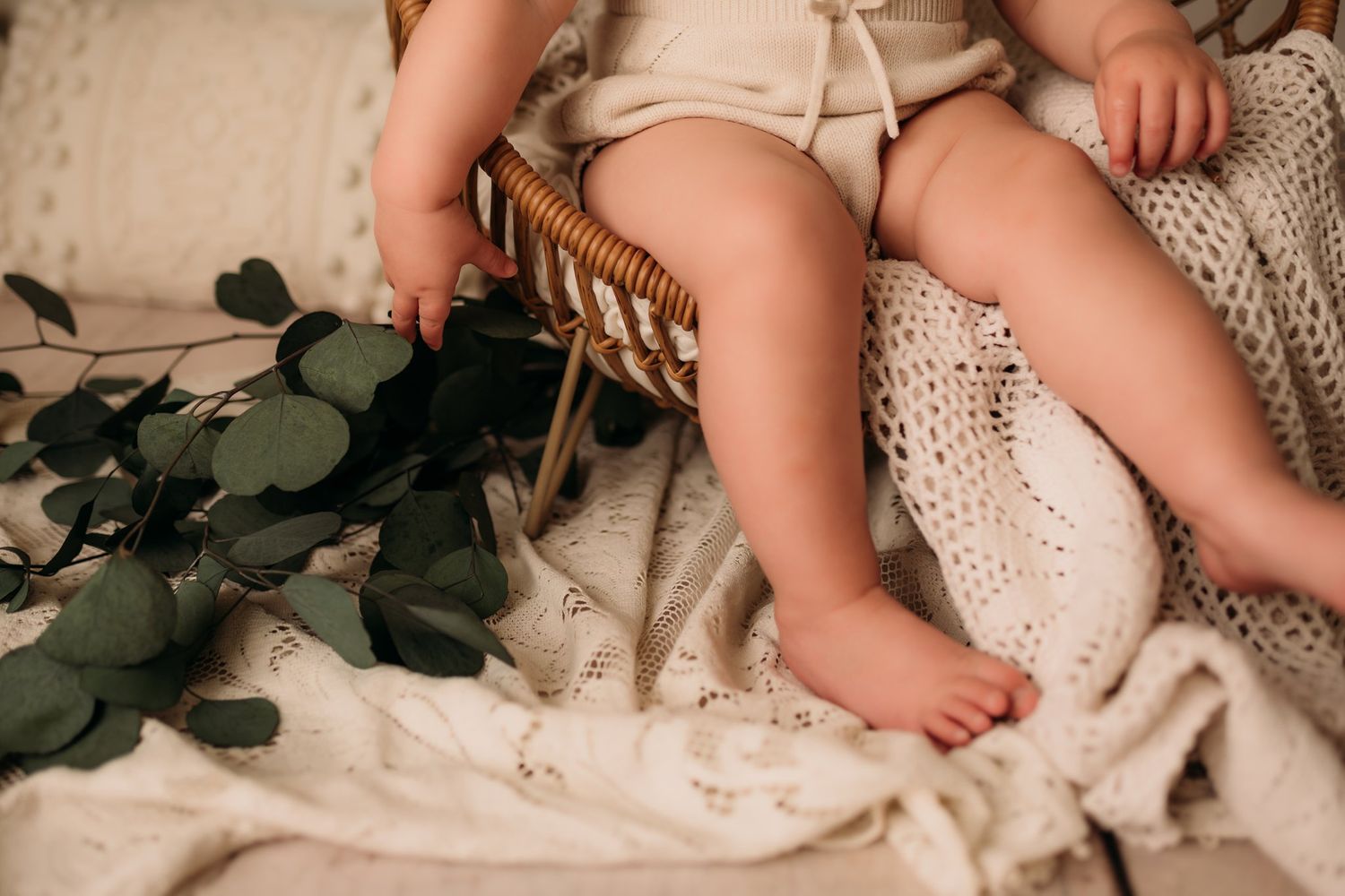 Close-up of tiny feet and cream romper against knit blanket and eucalyptus leaves.