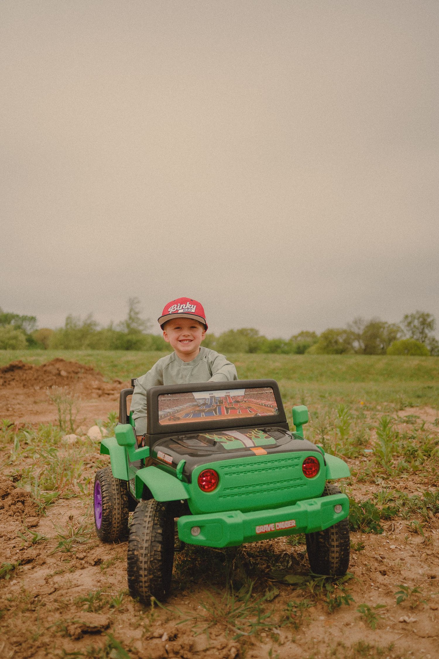 Smiling boy in red cap rides green Monster Truck toy jeep on muddy dirt field outdoors.