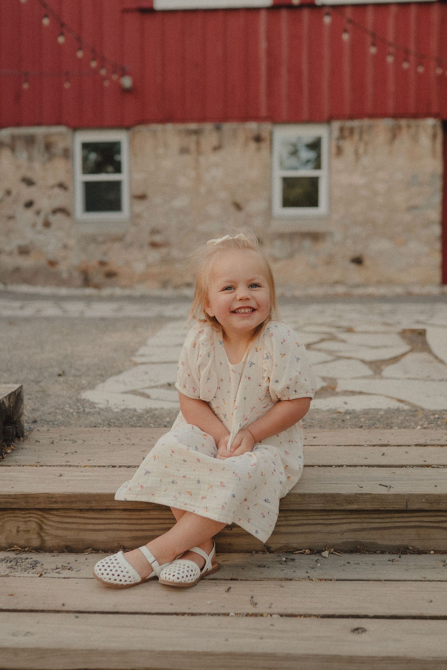 Adorable blonde toddler girl in cream floral dress smiling while seated on wooden steps near a red barn exterior.