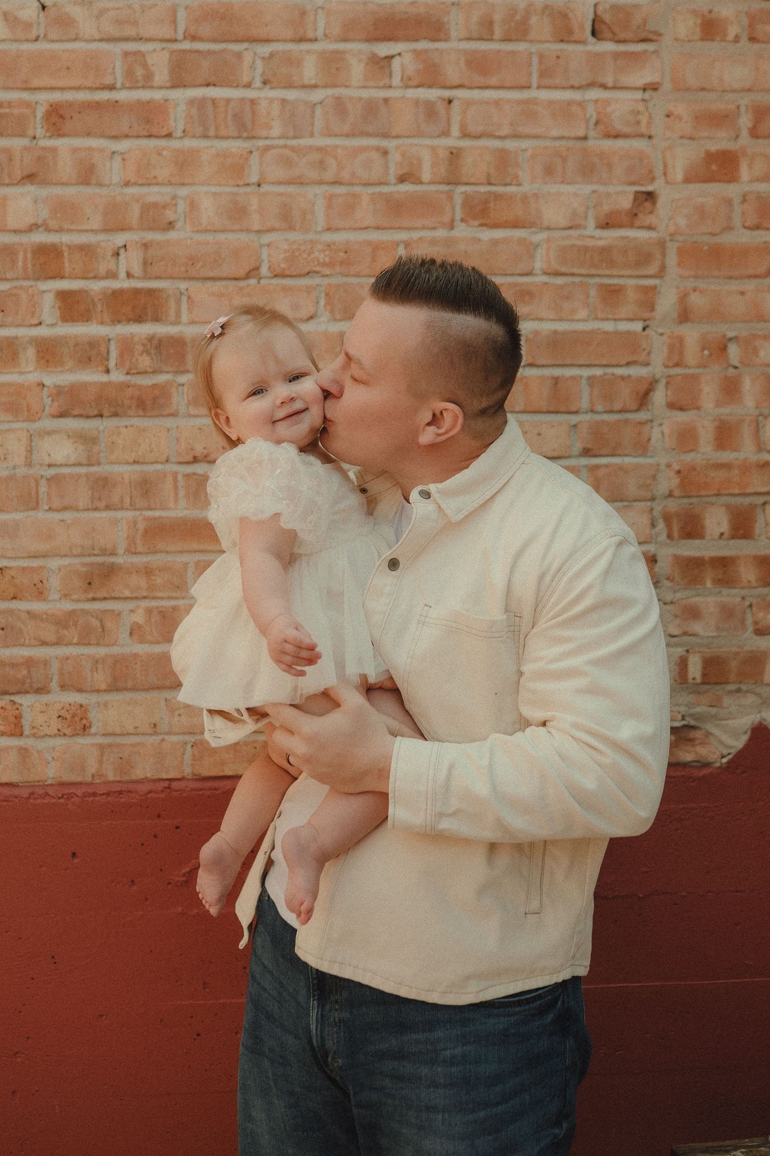 Father kissing his baby daughter on the cheek while holding her against a brick wall.