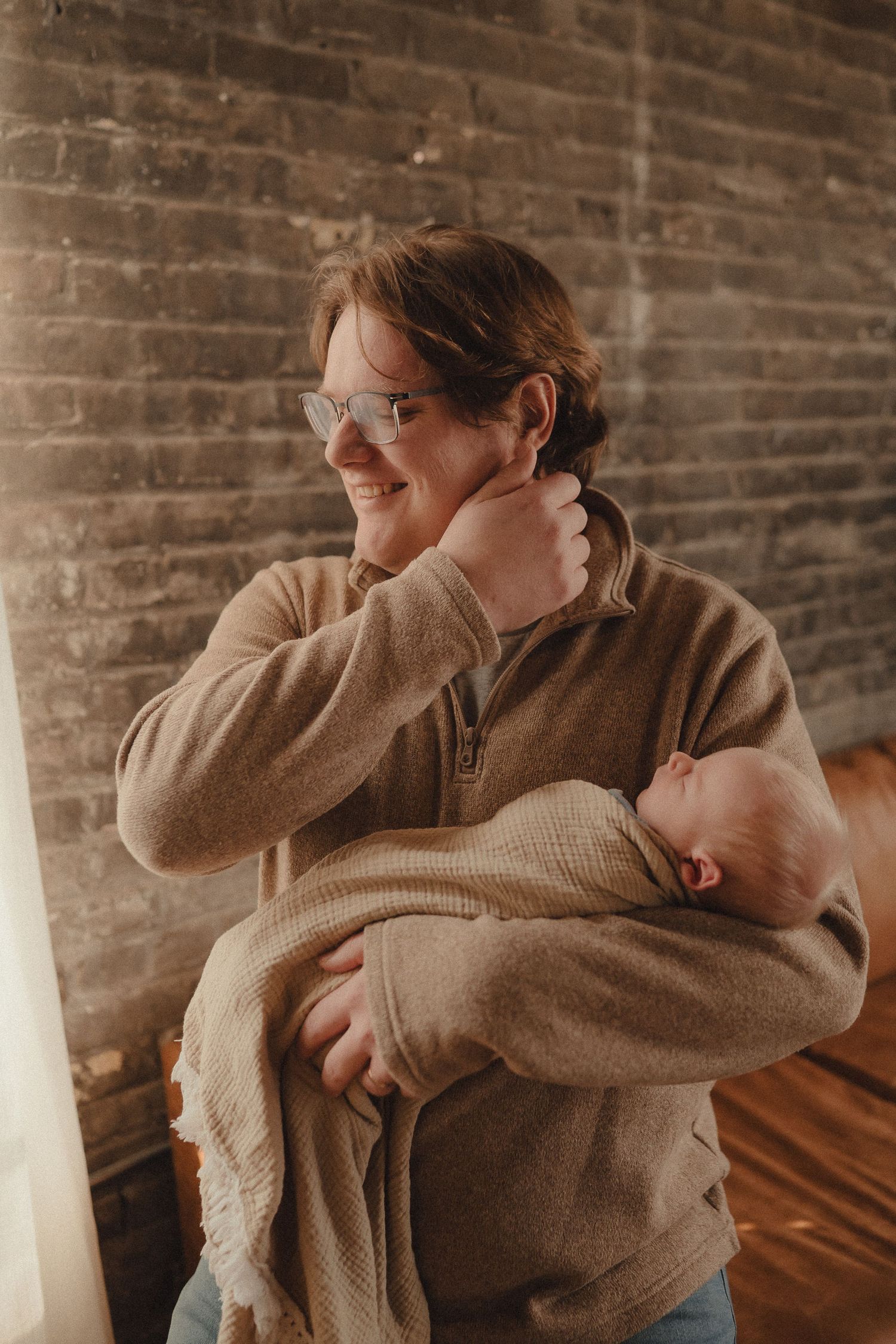 Smiling parent in tan sweater holding sleeping newborn baby in a warmly lit brick-walled room.