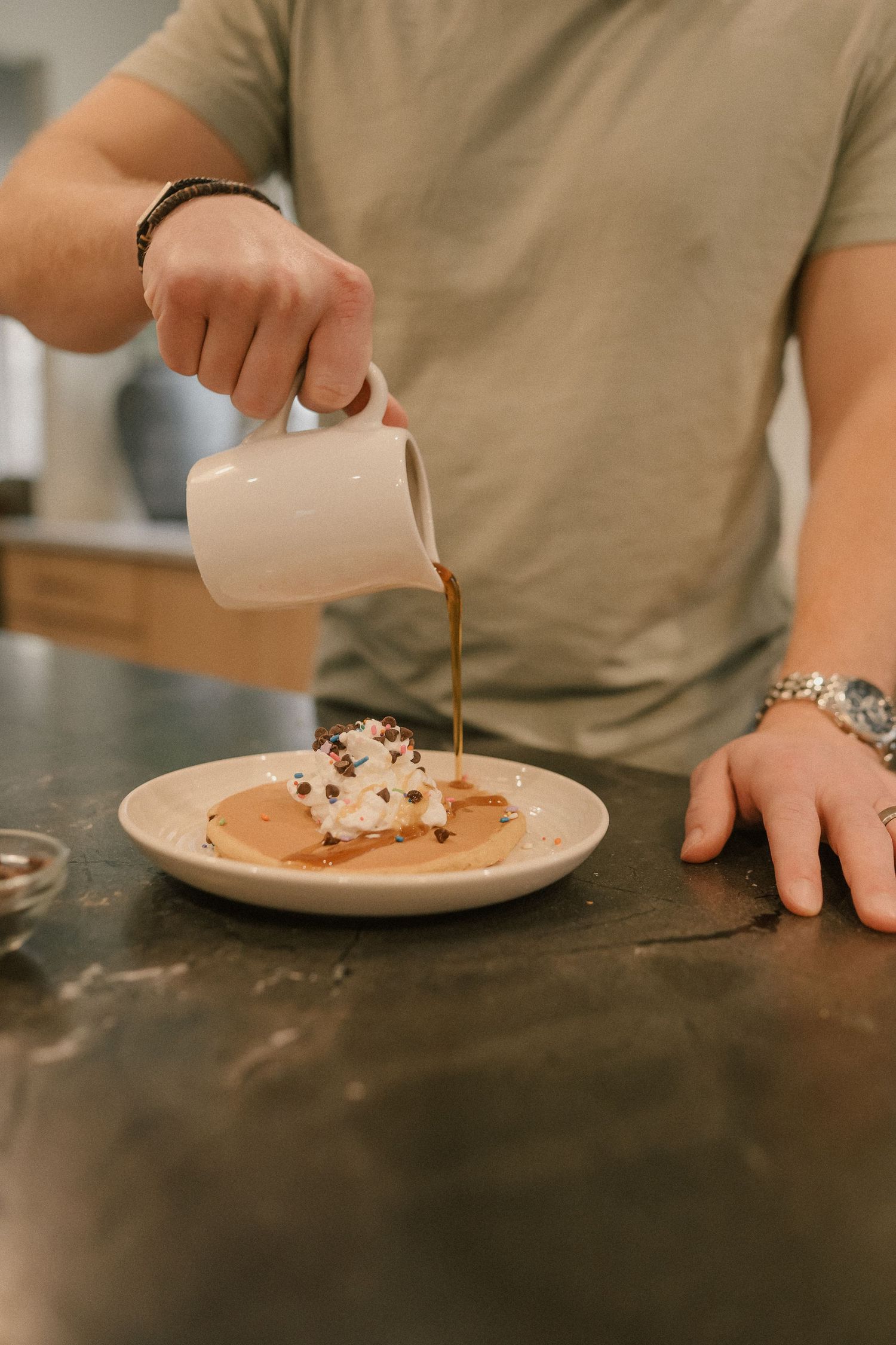 Man pouring chocolate syrup over whipped cream pancakes on a white plate at a kitchen counter.
