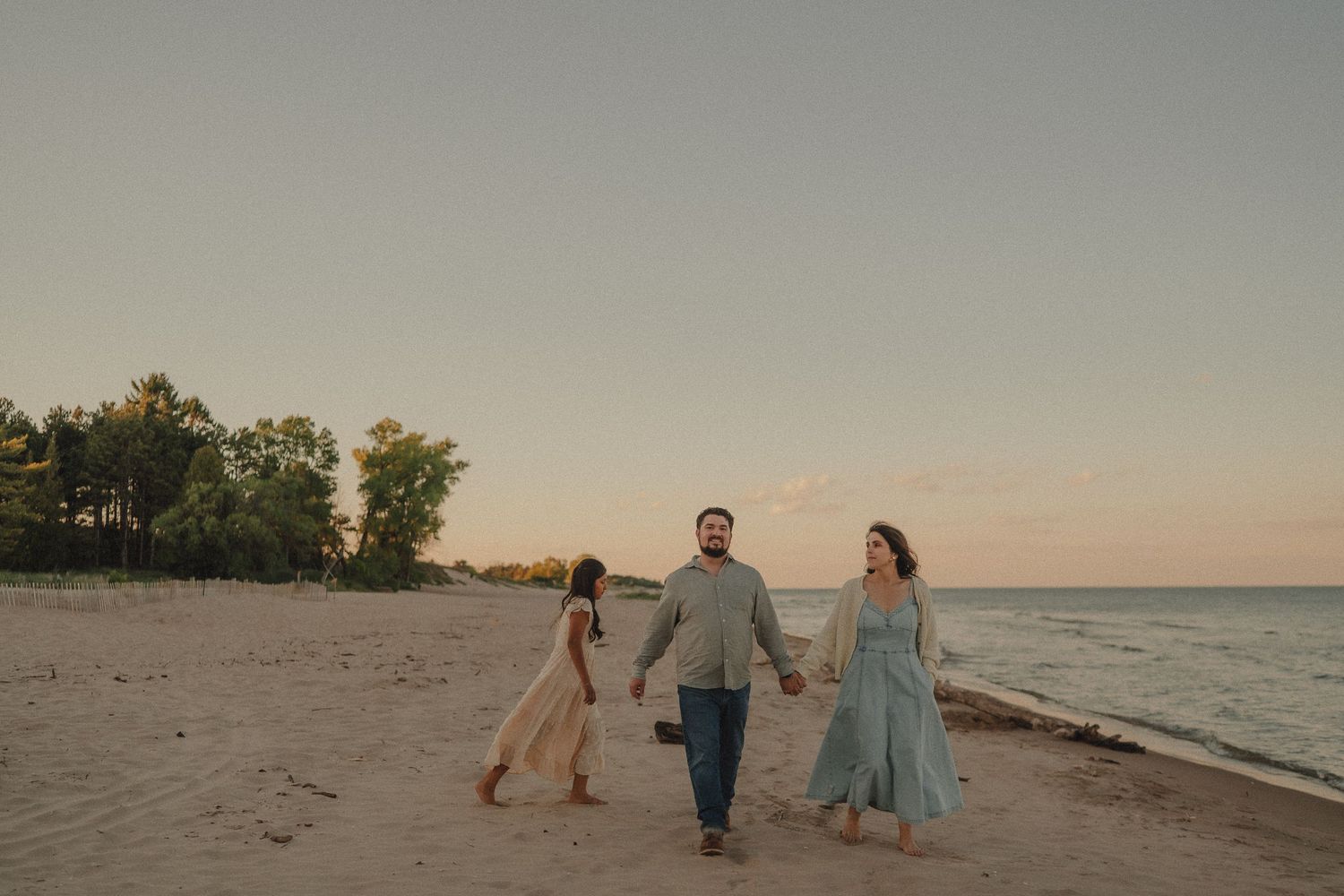 Family of three walks hand in hand along sandy beach at peaceful sunset.