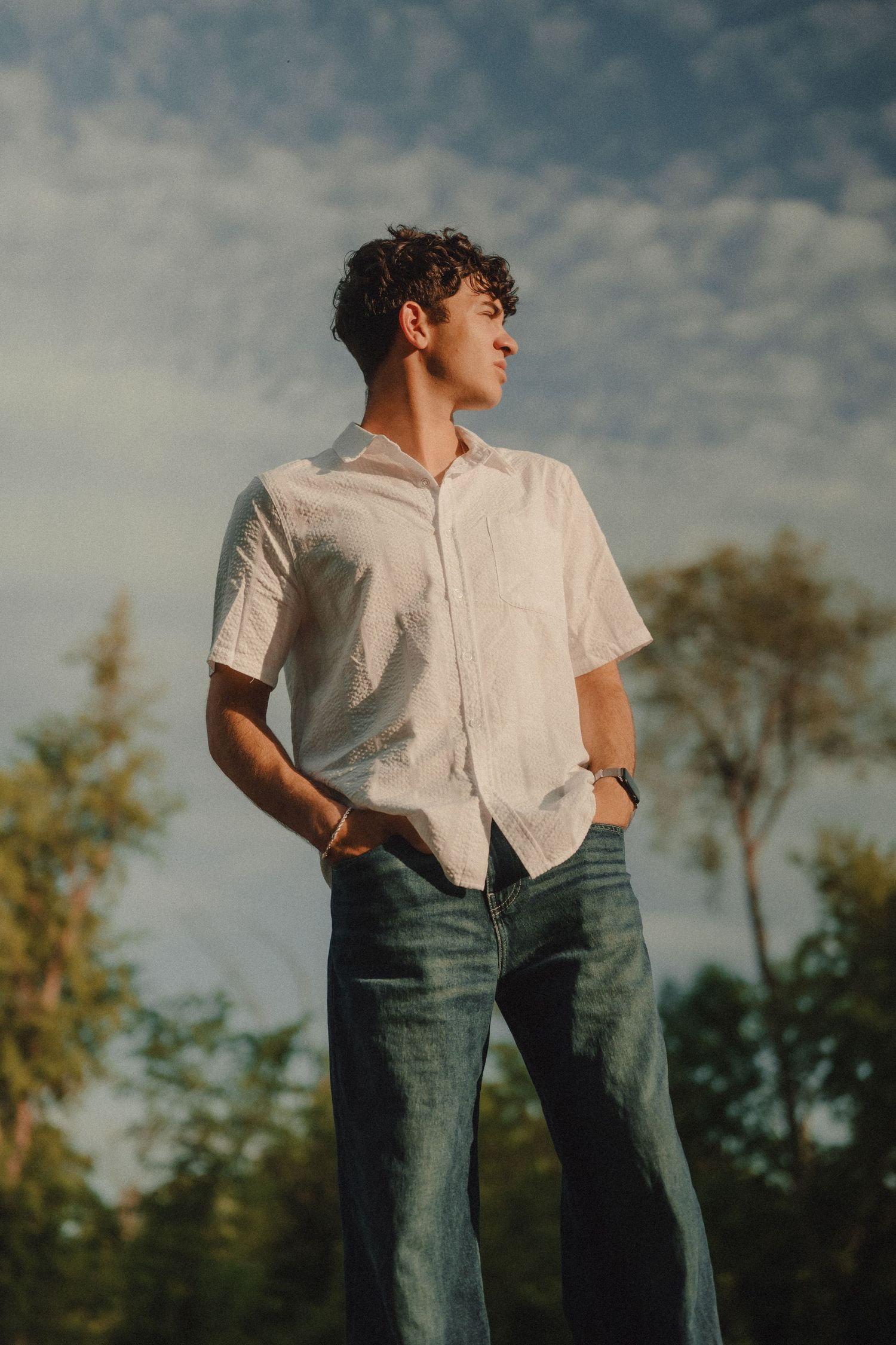 Young man in white shirt and jeans standing outdoors with trees and cloudy sky in background.