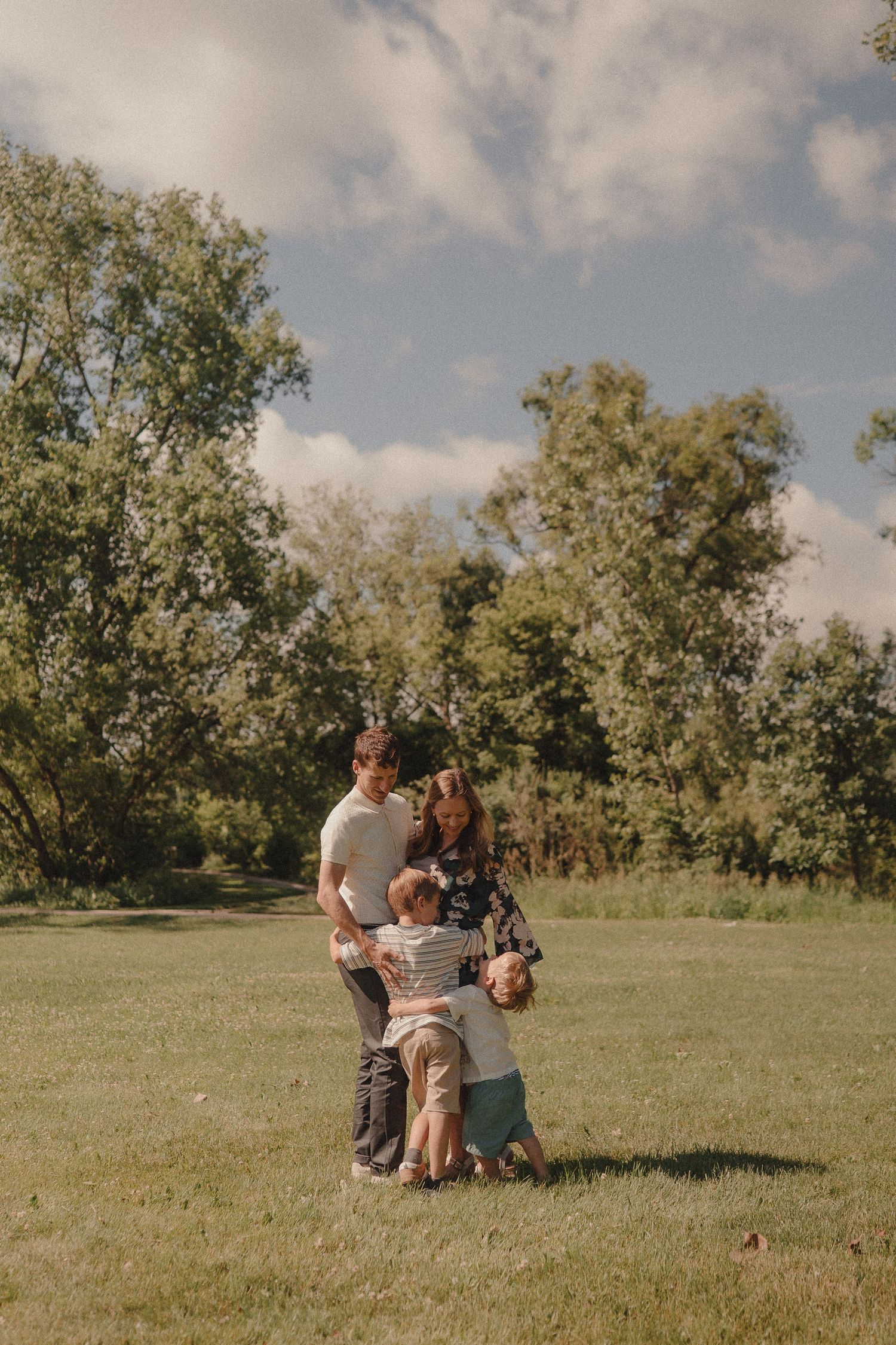 Family of four sharing a warm embrace in a lush green park under a partly cloudy sky.