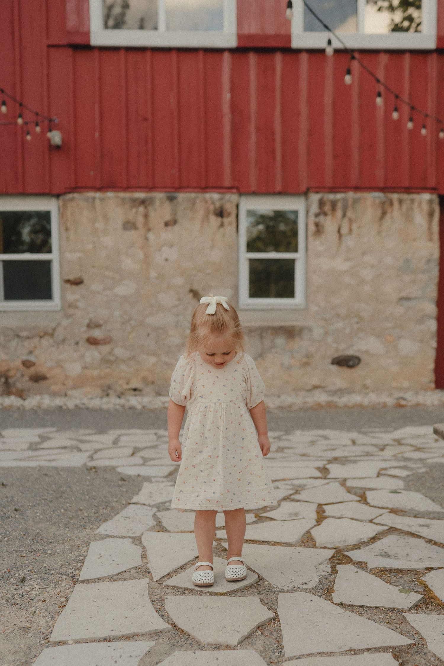 Toddler girl in a floral dress standing on a stone patio in front of a red barn.