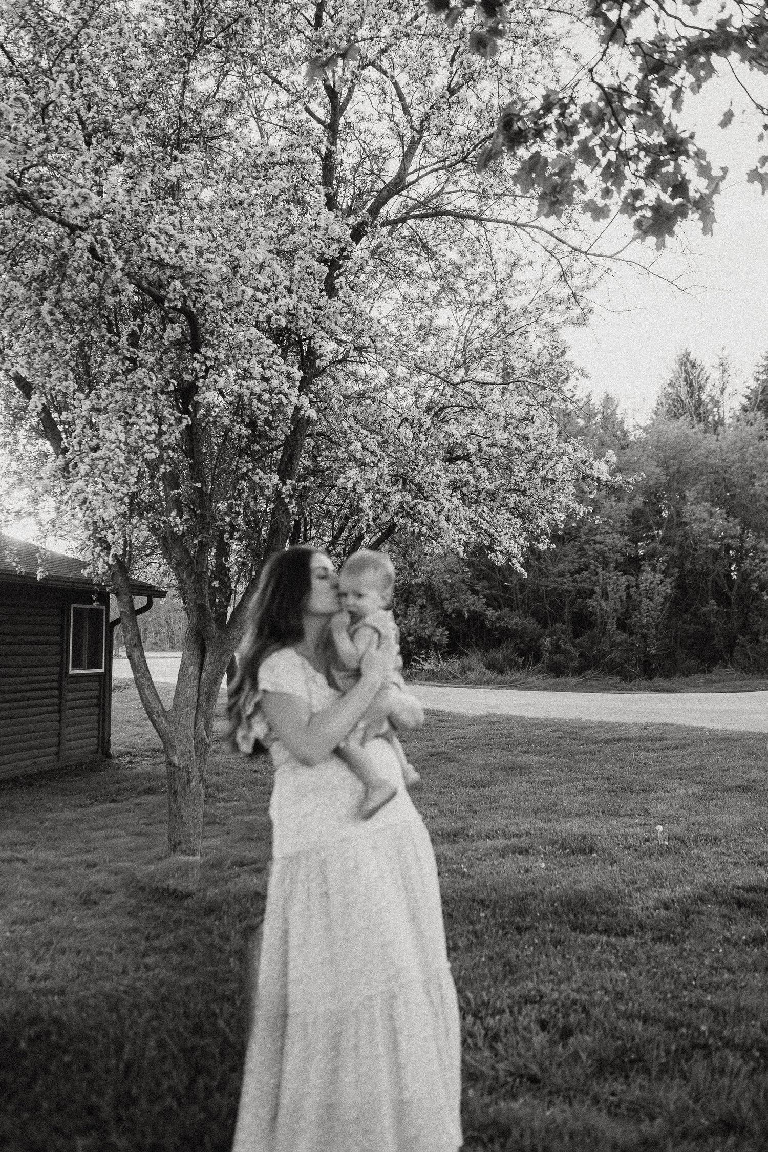 Black and white photo of mother holding baby under a blossoming tree in a peaceful yard.