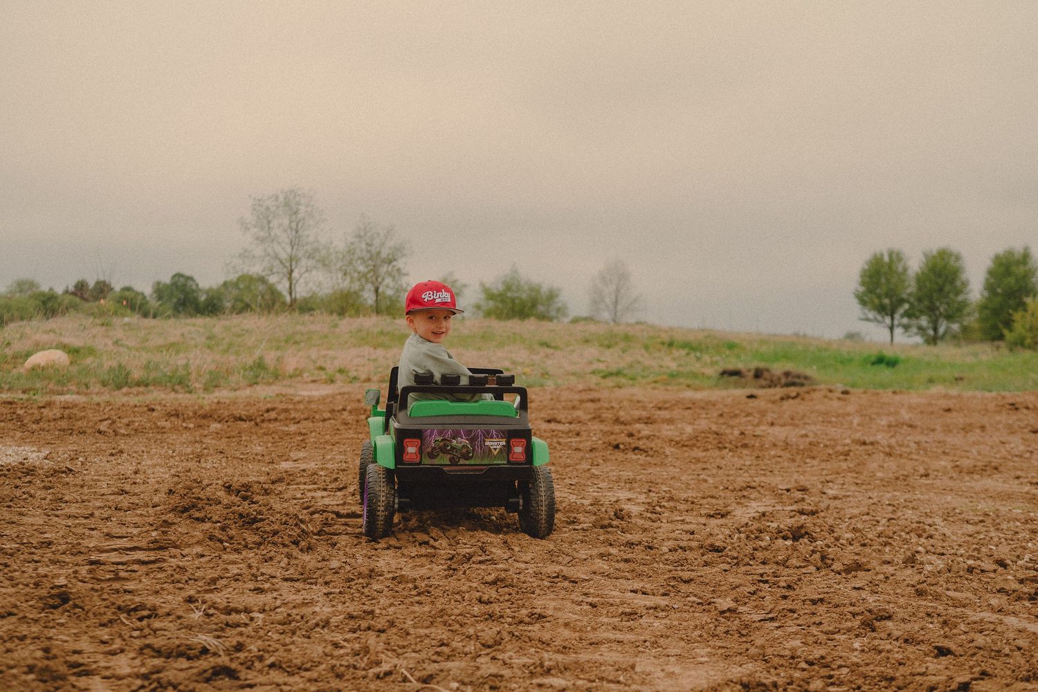 Child riding a green toy tractor across a plowed dirt field on a cloudy day.