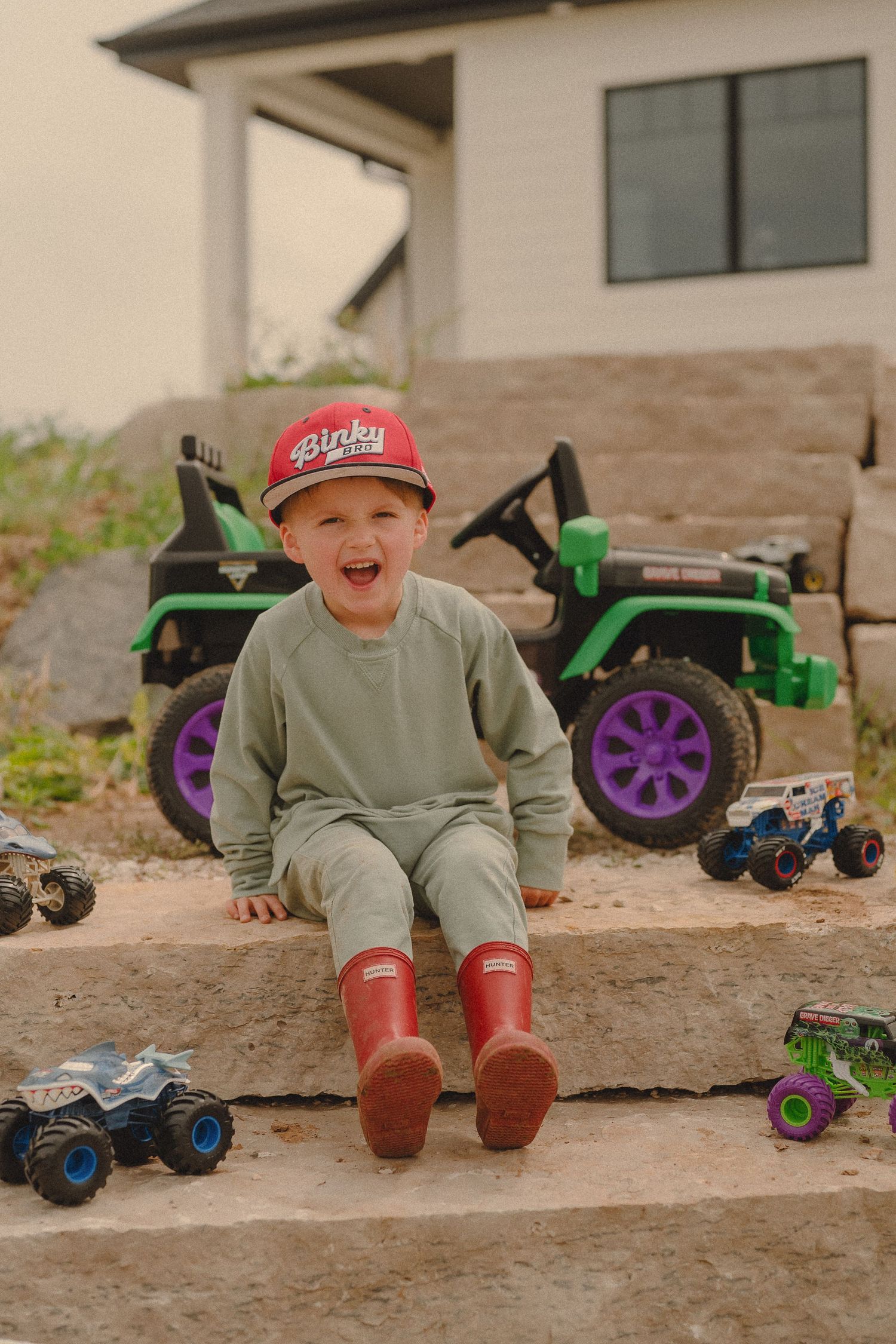 Laughing boy sitting on stone steps with monster trucks and green ride-on ATV.