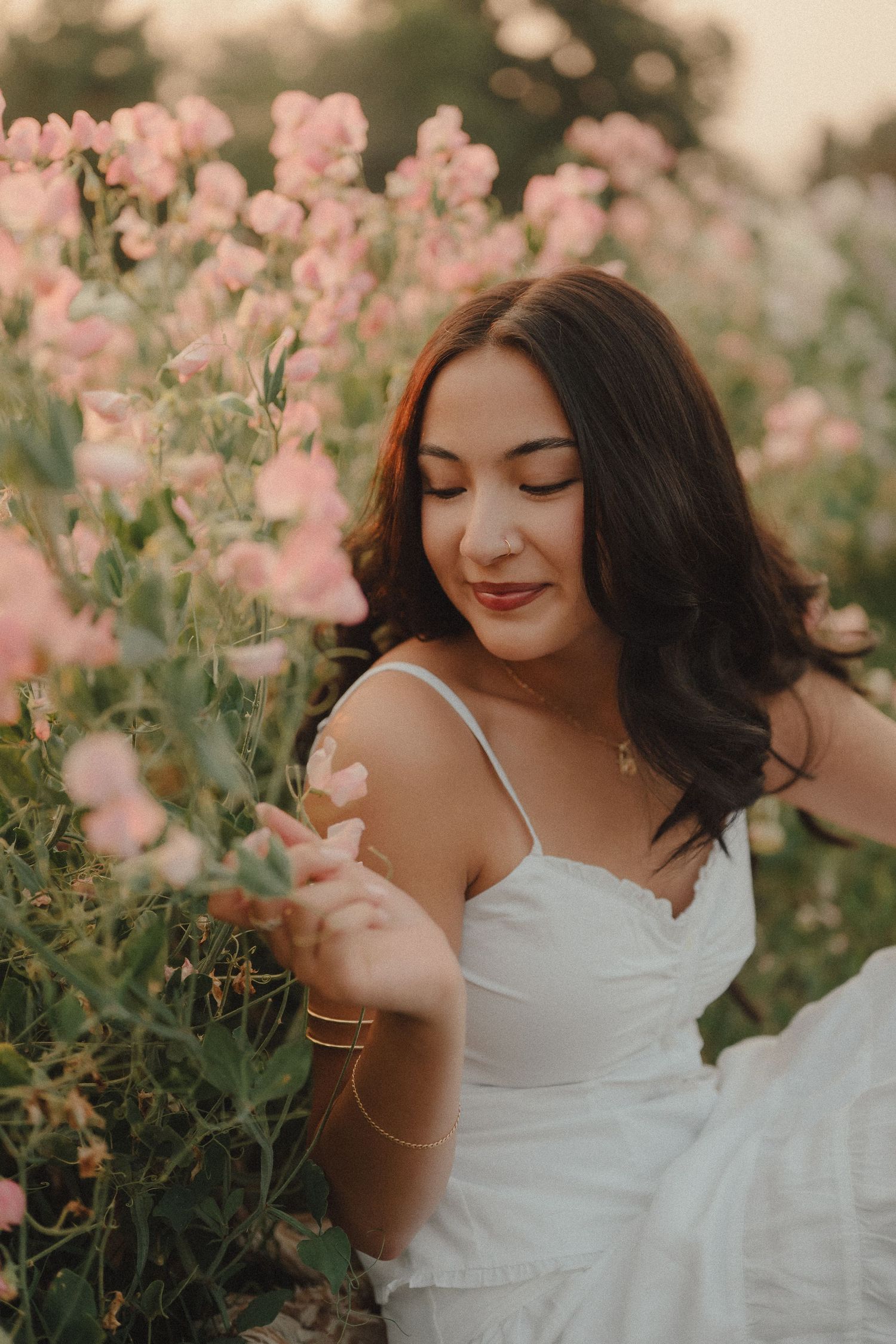 Young woman in white dress gently touching pink flowers in a blooming garden at dusk.
