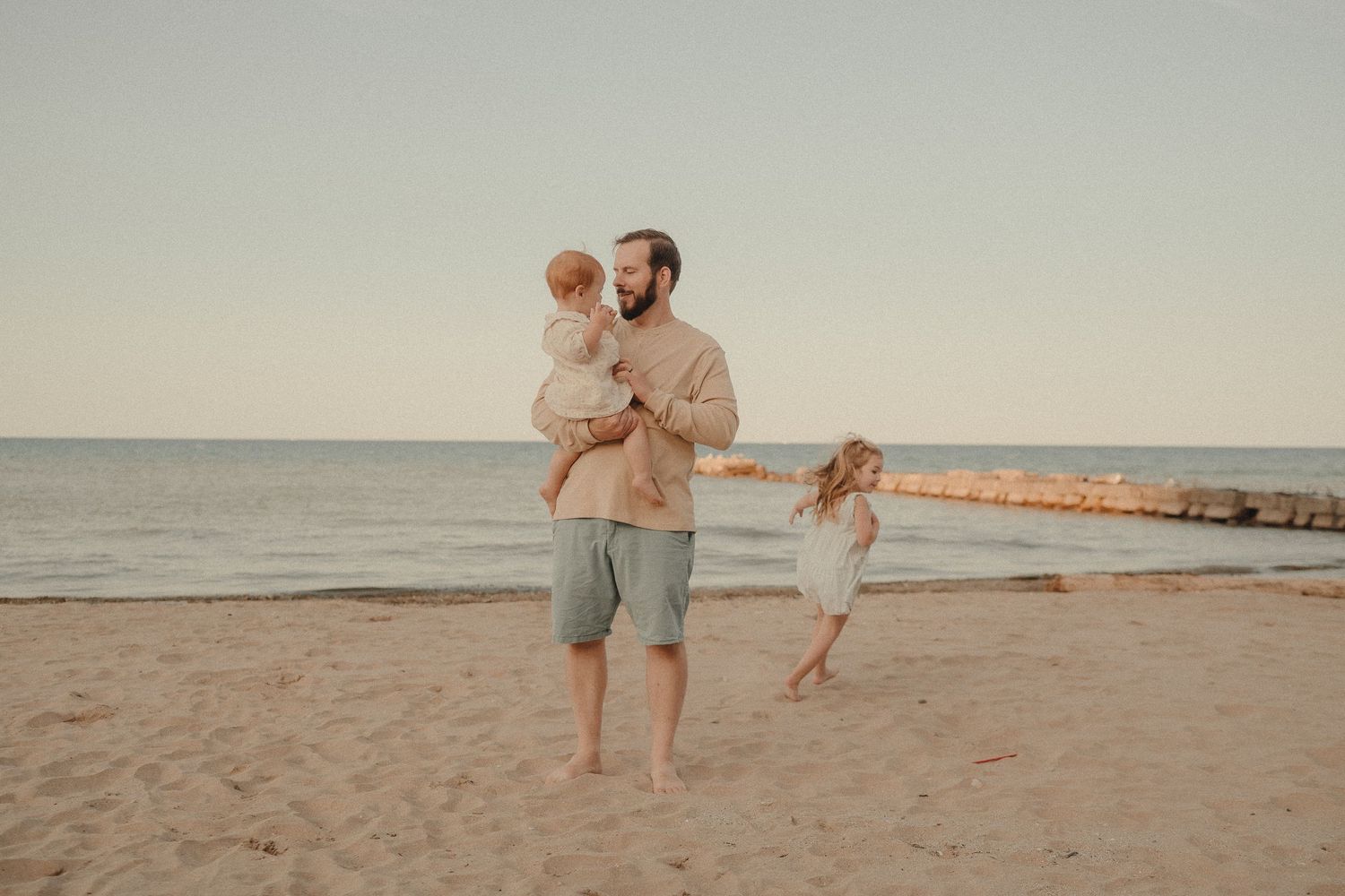 Father holding baby while young daughter runs toward him on sunny sandy beach with ocean backdrop.