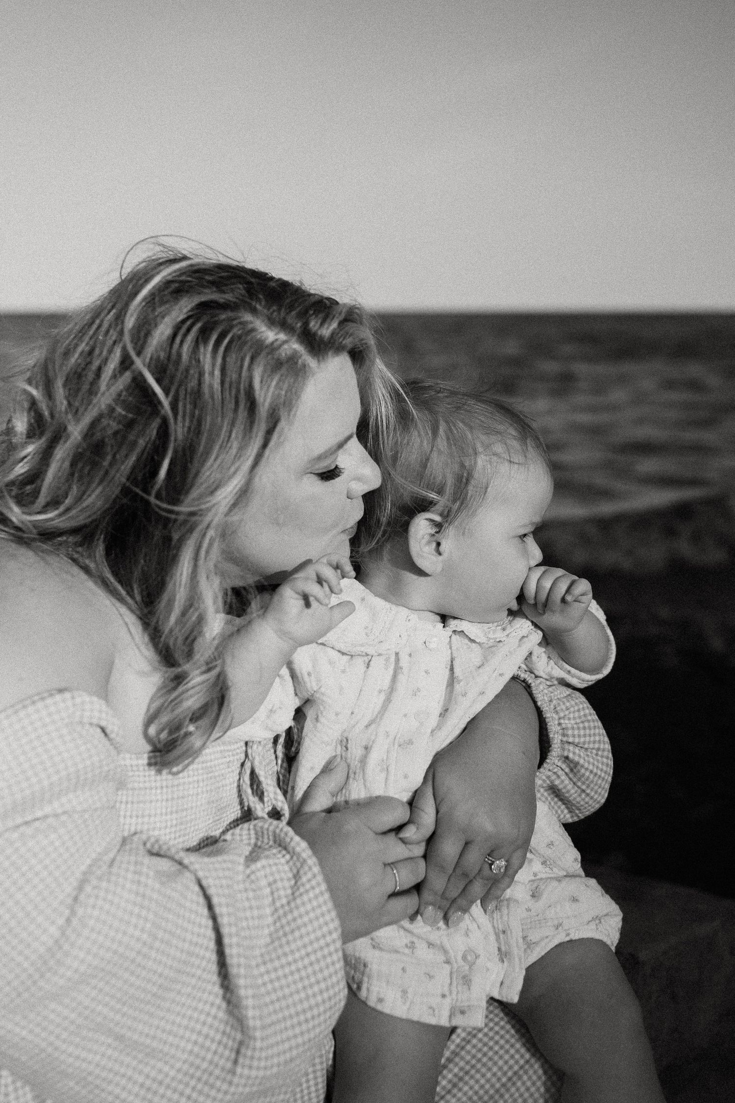 Black and white portrait of blonde mother tenderly holding and gazing at her baby by the ocean.