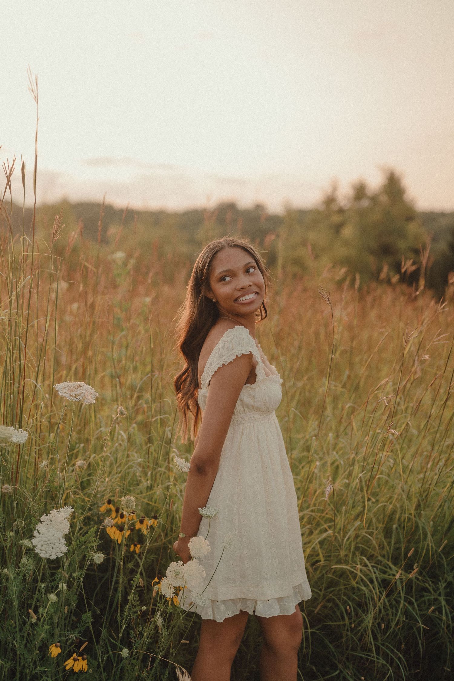 Smiling young woman in white lace dress holds wildflowers in sunset meadow.