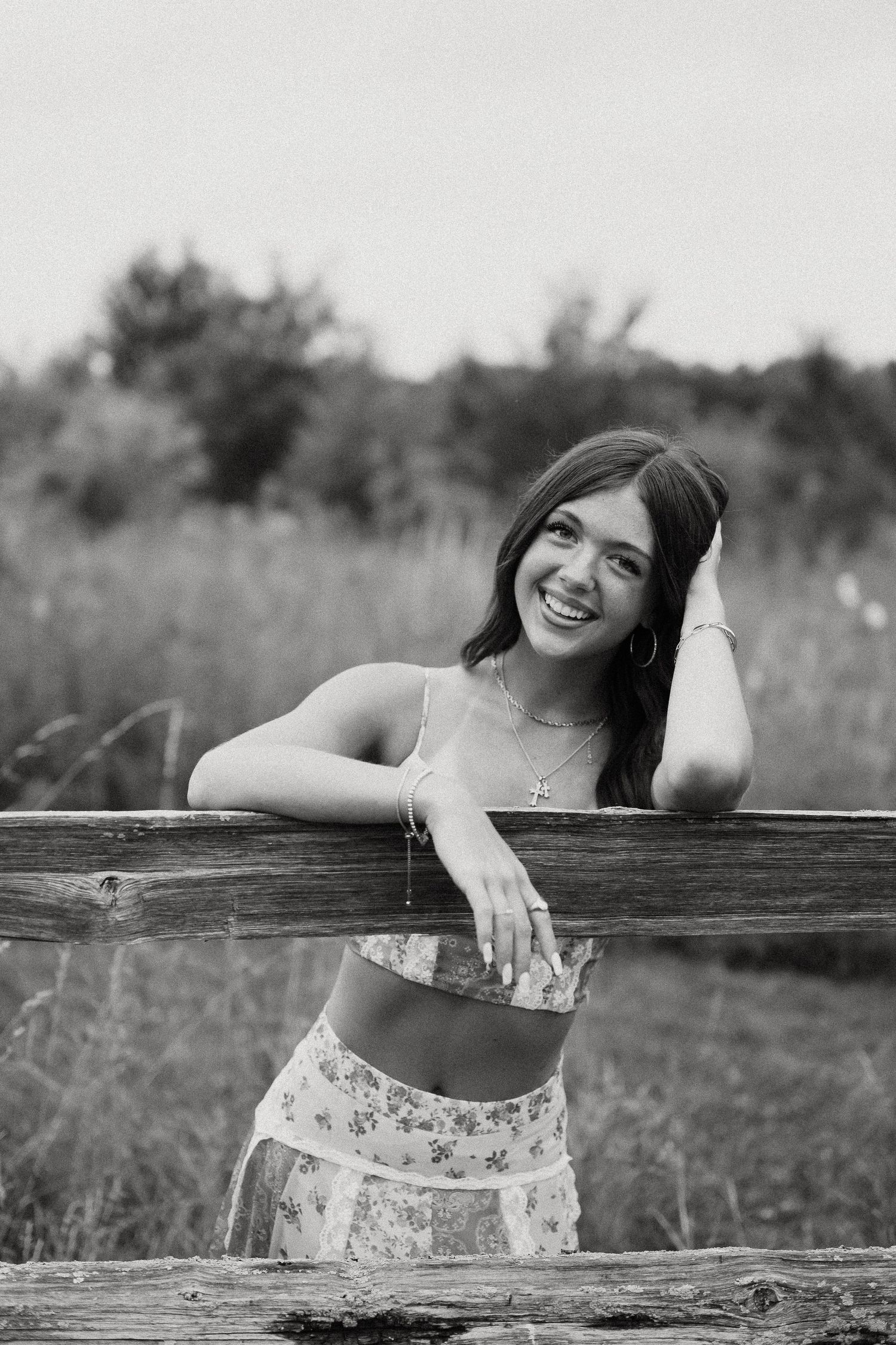 Black and white photo of a smiling young woman leaning on a rustic wooden fence in a field.