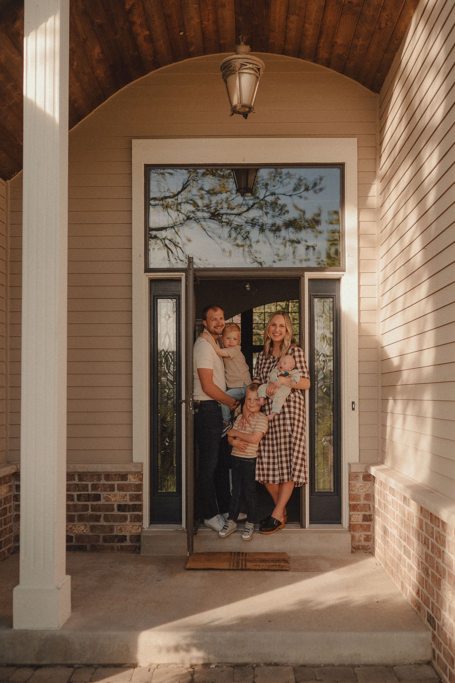 Young family of four posing at their front door under a wooden arched porch with brick accents in warm evening light.