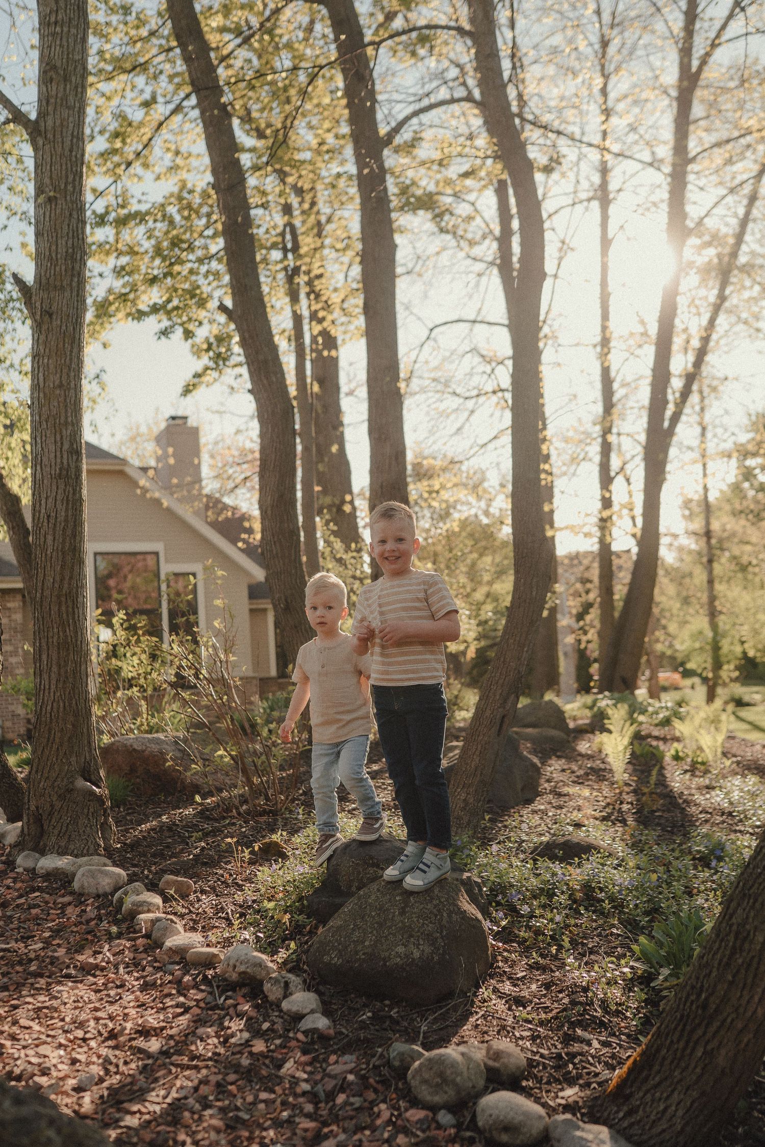 Two young boys standing on a large rock in a sunlit backyard surrounded by trees.