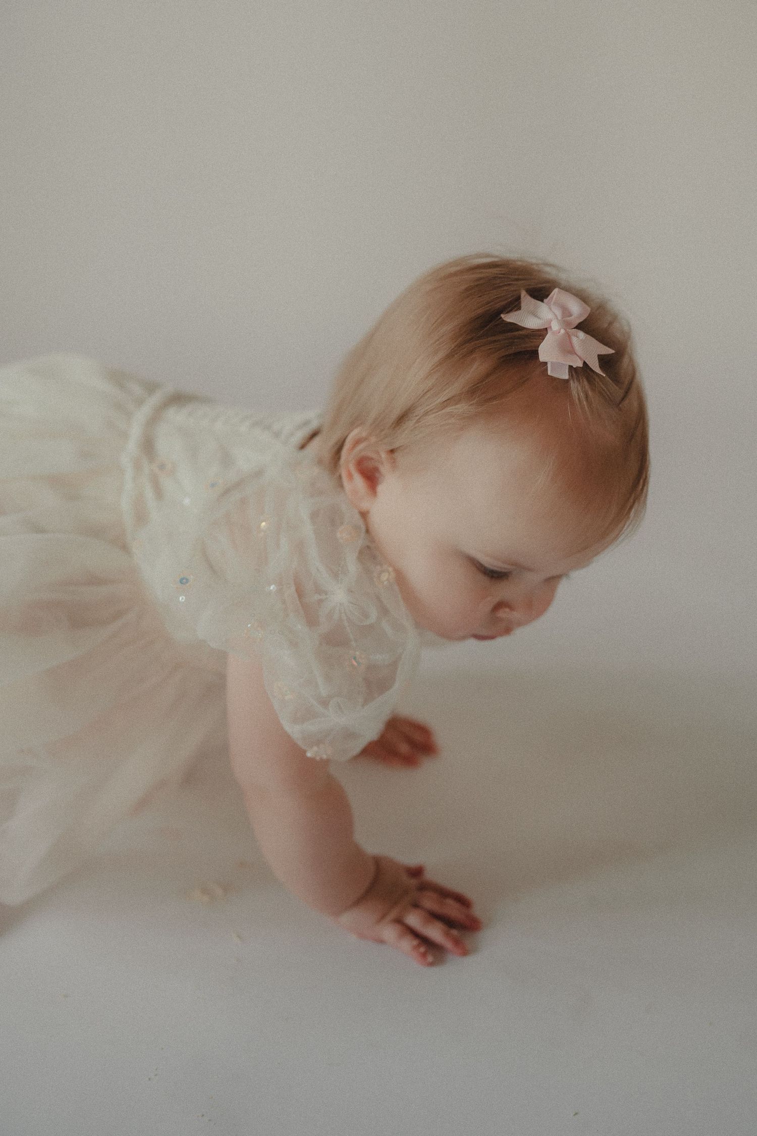 A cute baby girl in a cream lace dress with a pink bow crawls on a white surface.