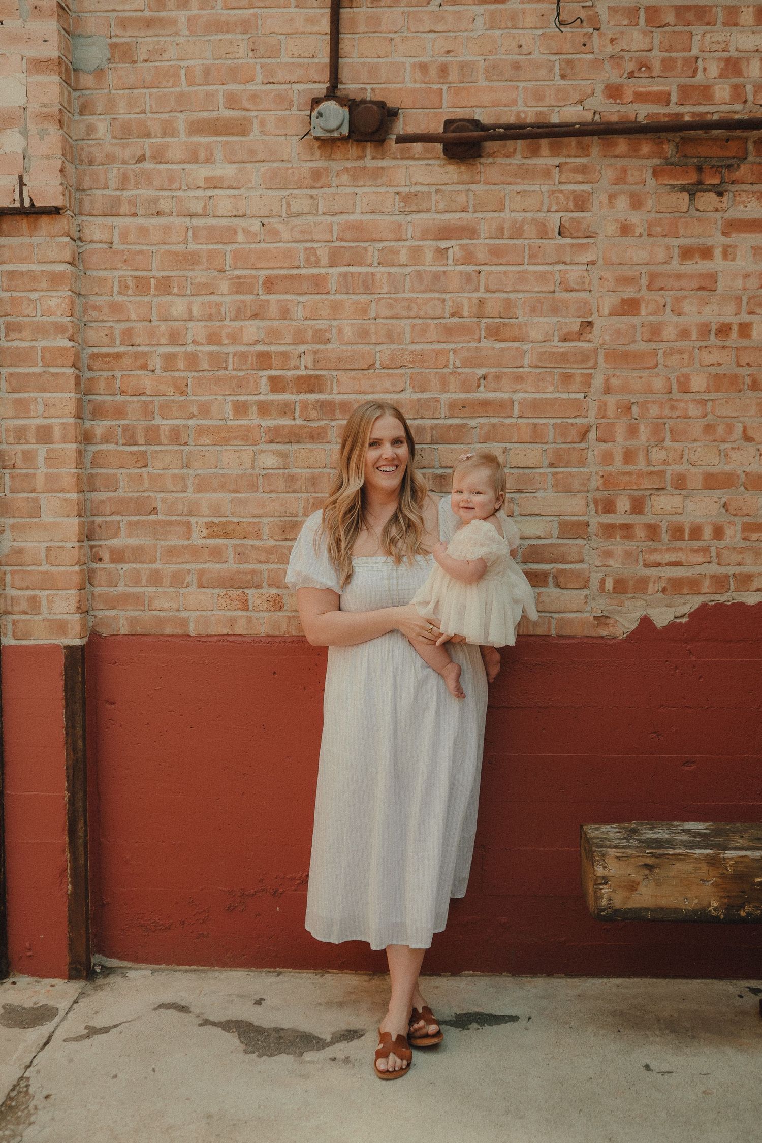 A blonde mother in a white midi dress holds her baby girl while posing in front of a rustic brick wall.
