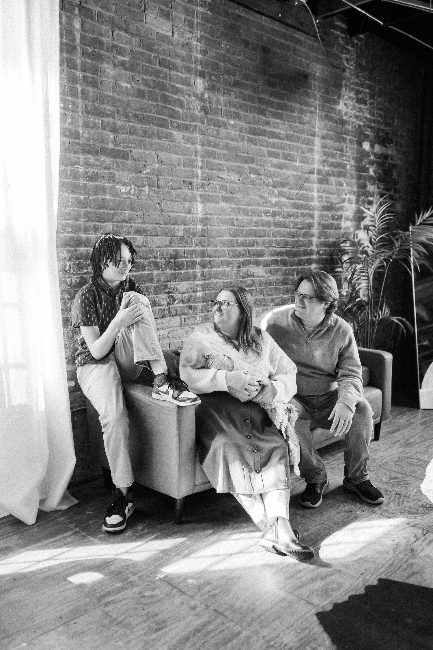 Black and white photo of three people sitting together on chairs near a brick wall, with one holding a newborn baby.