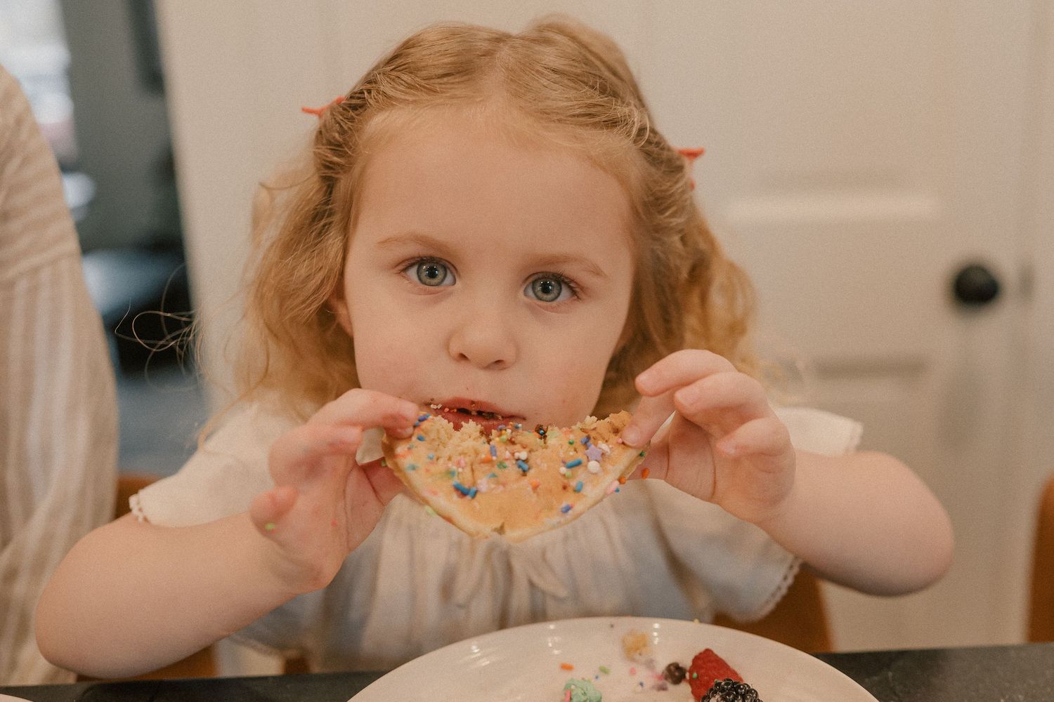 Young curly-haired girl eating a colorful sprinkle-covered pancake slice with both hands.