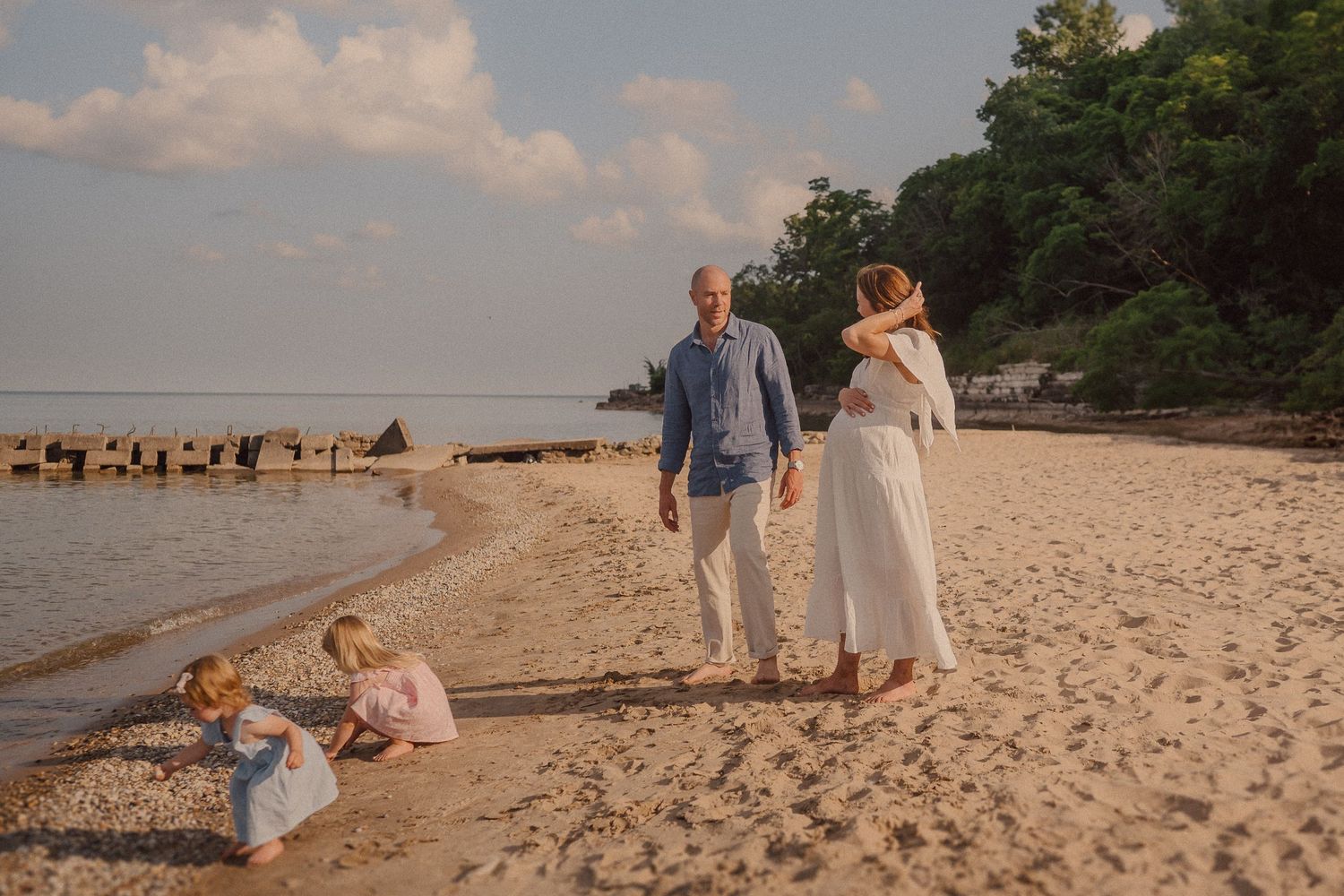 A family walks along a sandy beach at sunset with wooden pilings visible in the water.
