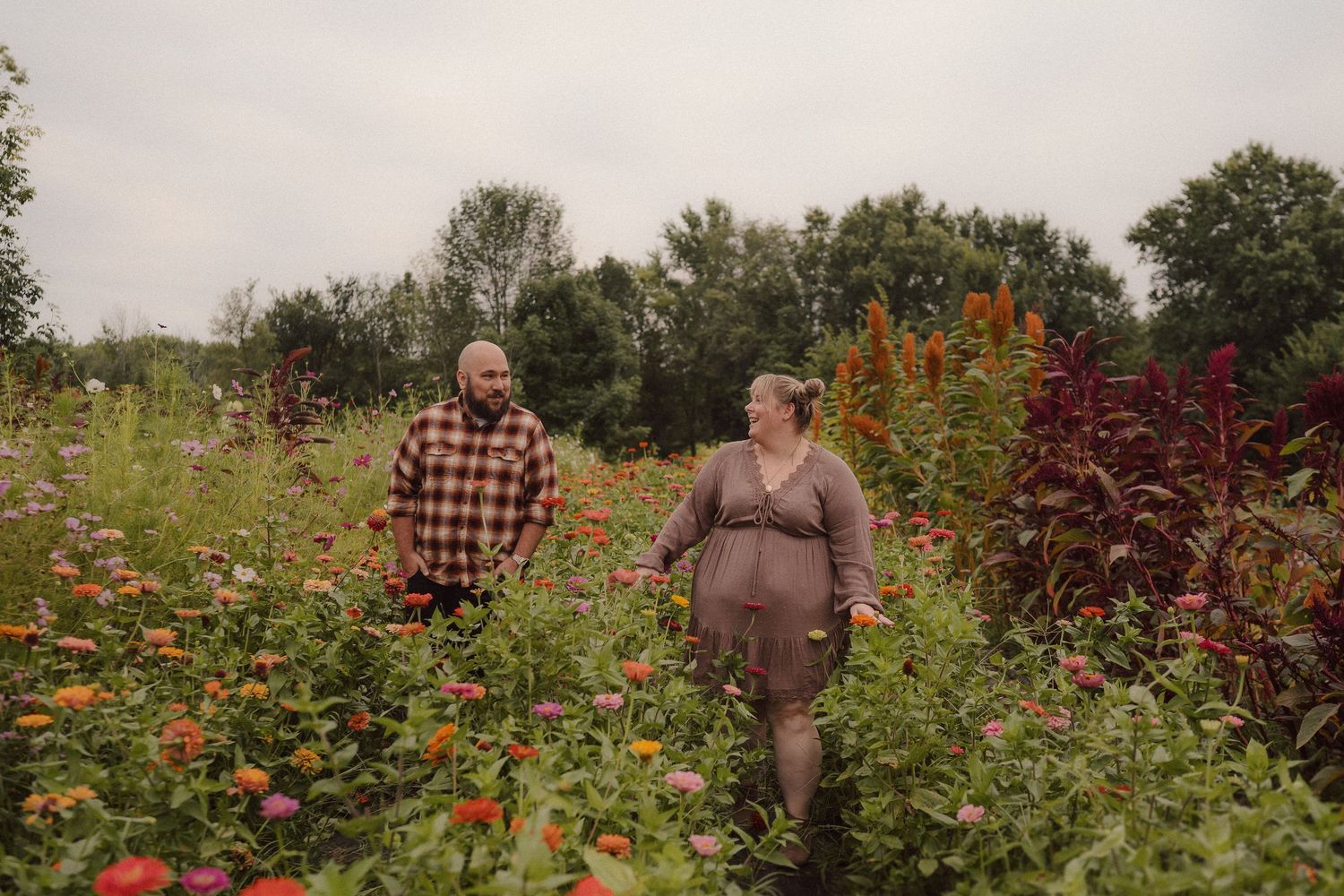Two people stand together in a vibrant garden filled with bright flowers and lush vegetation on a cloudy day.