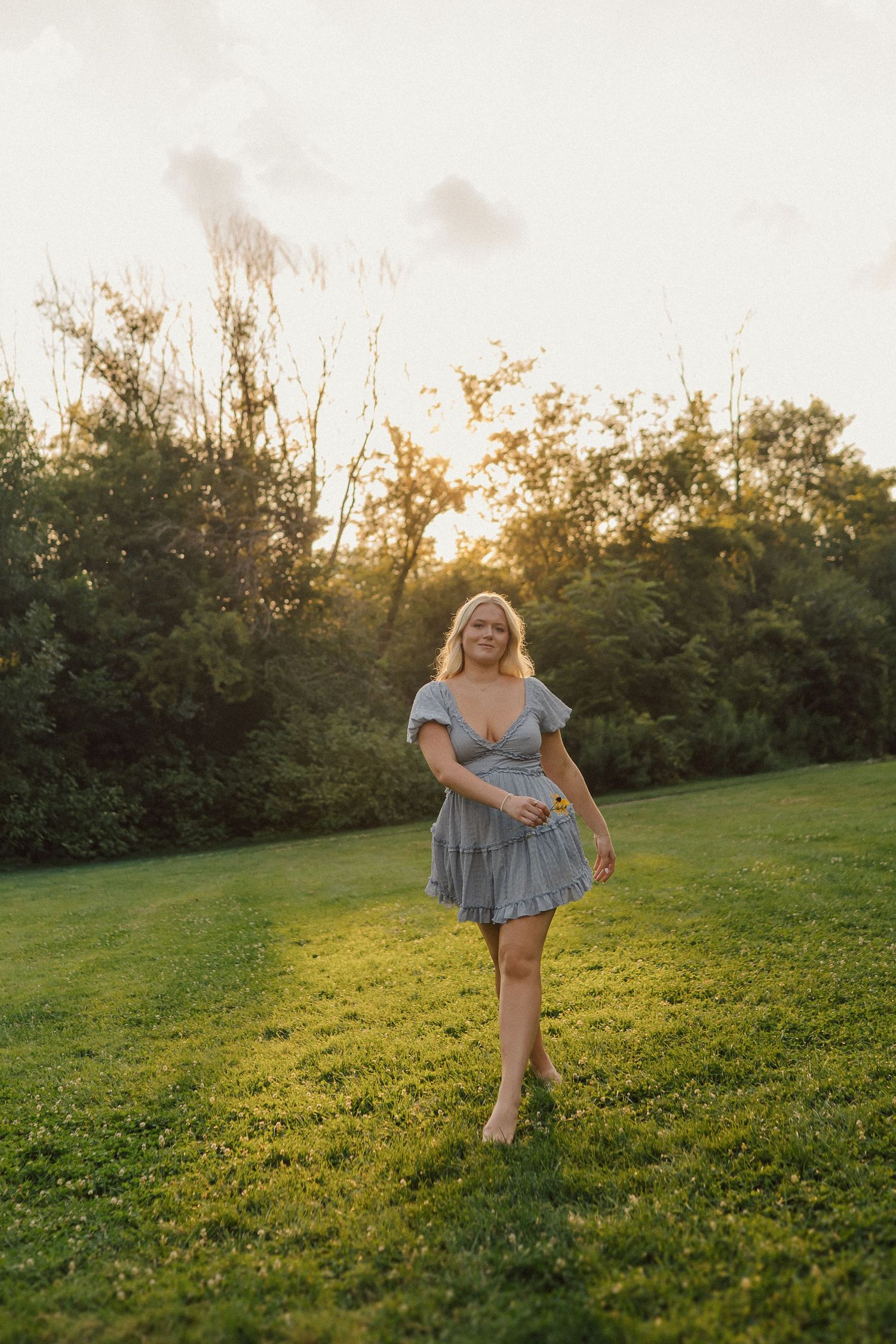 Person in a light blue dress spins playfully on a grassy hillside during golden hour sunset.