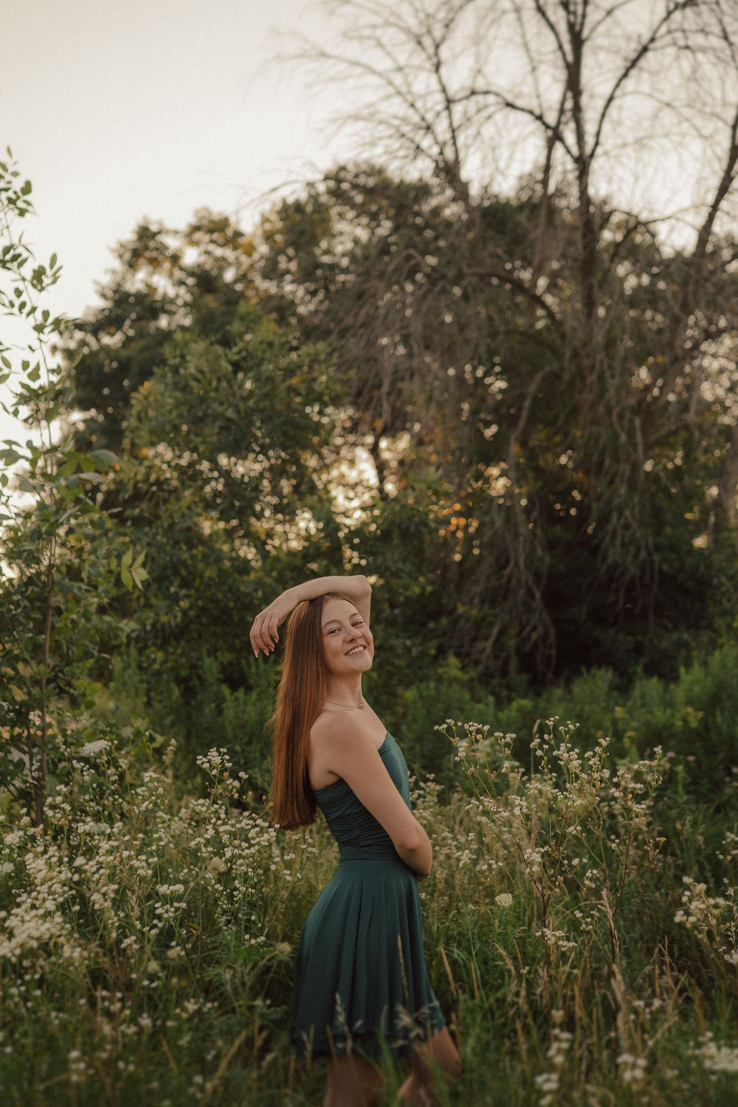 Person in emerald dress dances through a sunlit meadow with wildflowers and trees at golden hour.