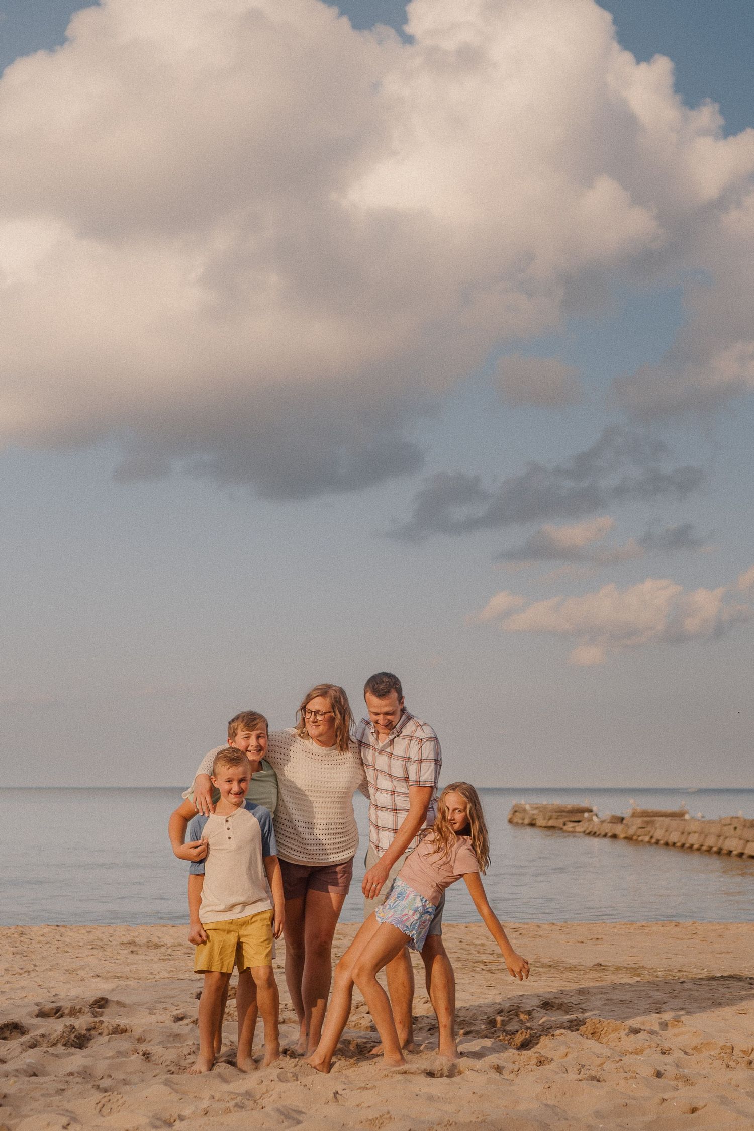 A family of five poses for a portrait on a sandy beach at sunset with fluffy clouds in the background.