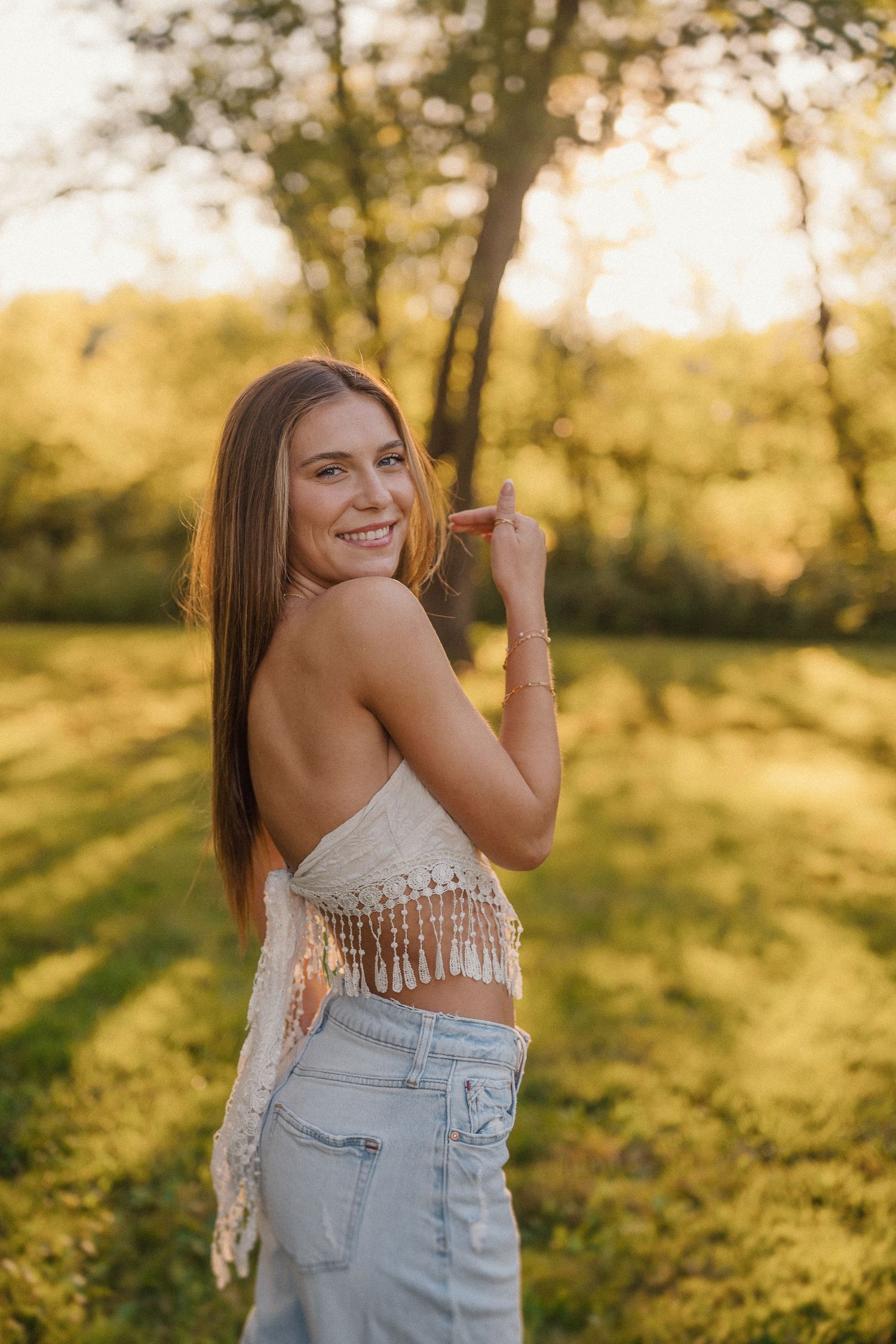 Woman in fringe crop top and jeans poses playfully in golden sunset light.