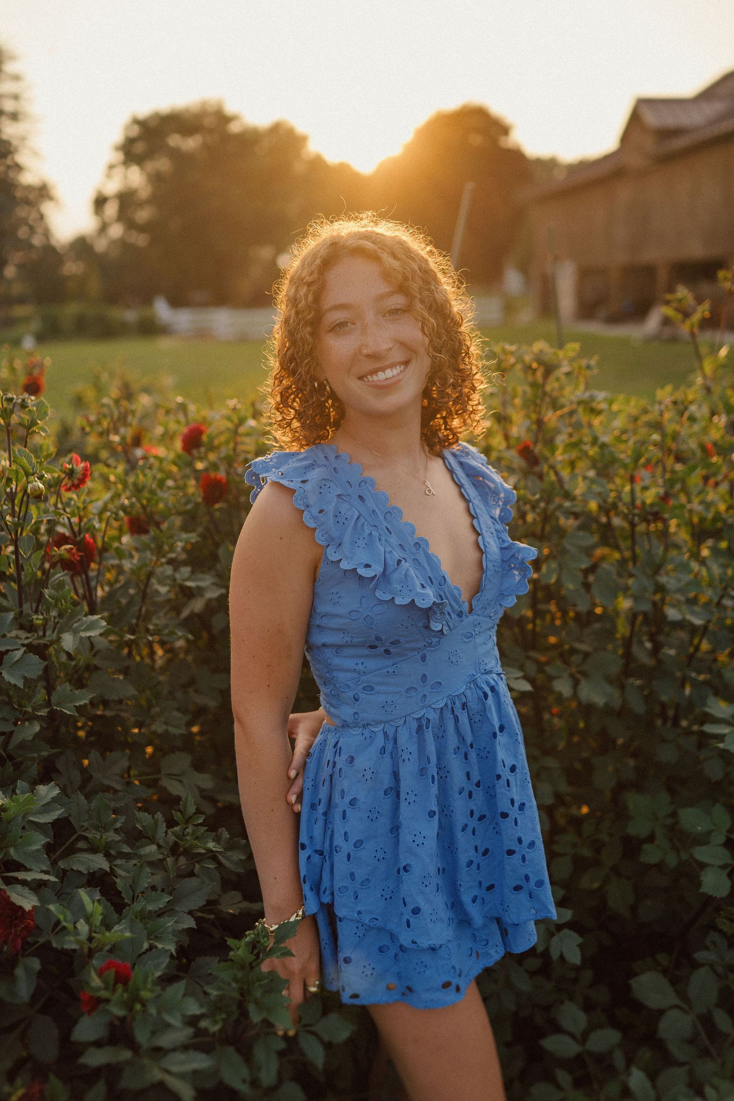 Sun sets behind a rose garden as a person in a blue eyelet top smiles warmly during a summer evening photoshoot.