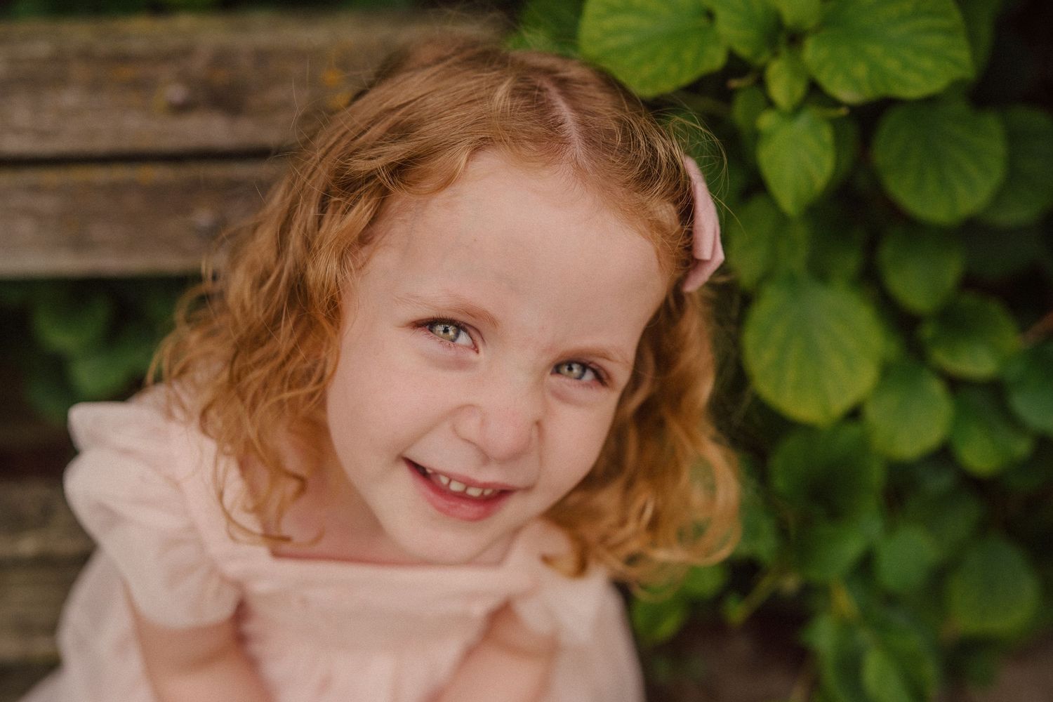 Young child with curly hair smiling at camera in pink dress with green foliage in background.