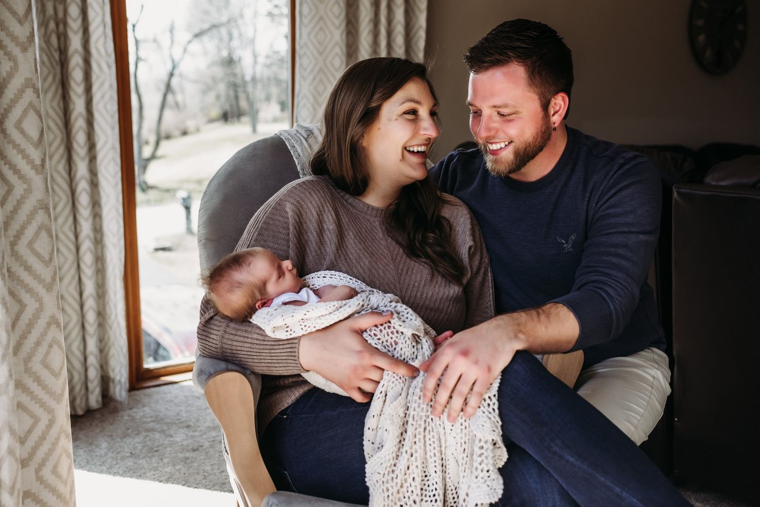 Intimate family moment by window as parents share joy with newborn wrapped in knitted blanket.