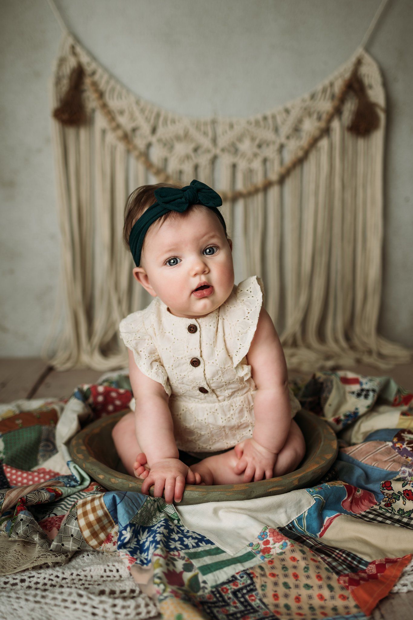 Baby in cream ruffled romper with teal headband sits in wooden bowl on vintage quilt.